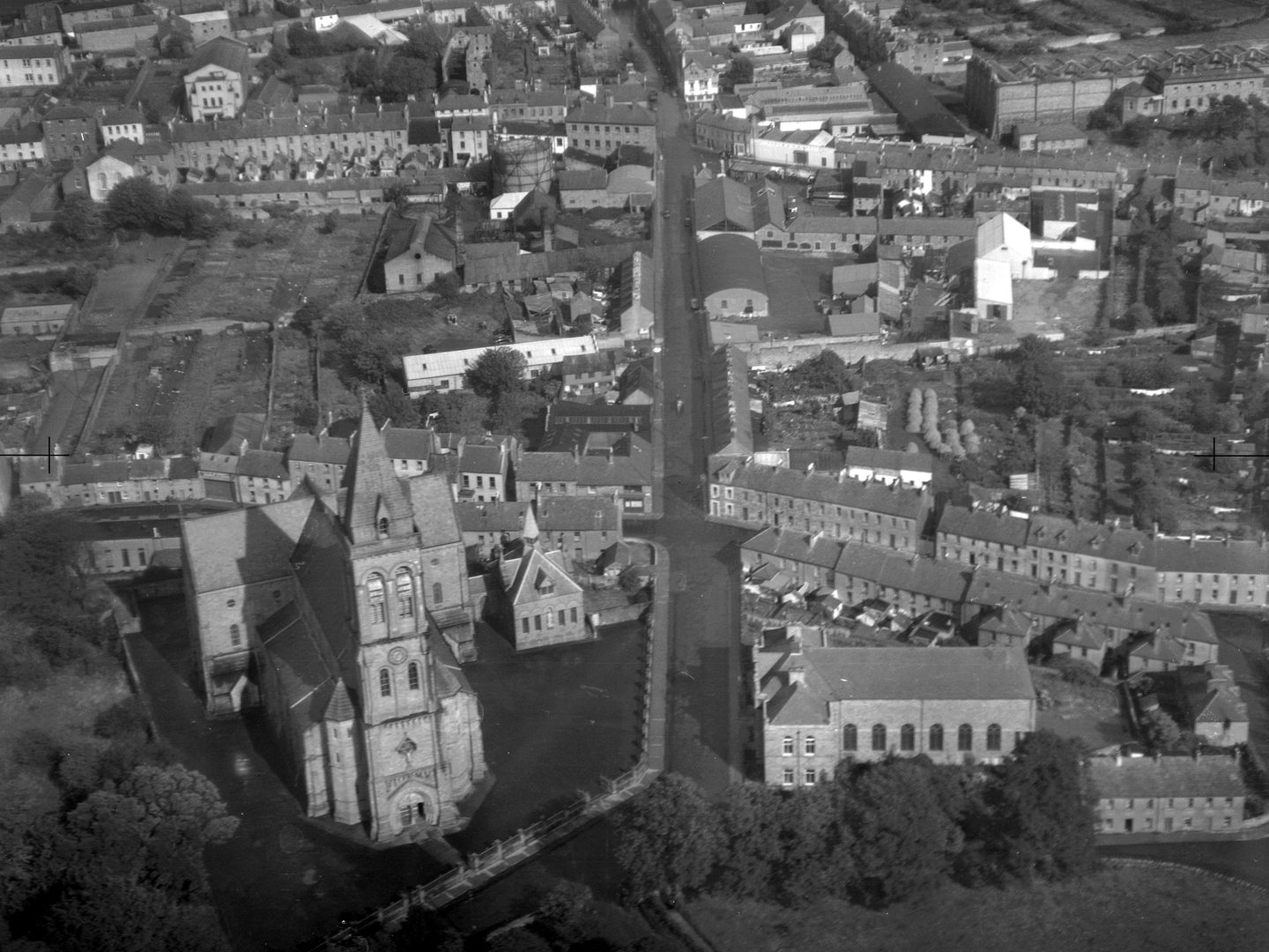 A historic black and white oblique vertical aerial photo of a town. In the foreground is a cathedral with a tower and short transepts.