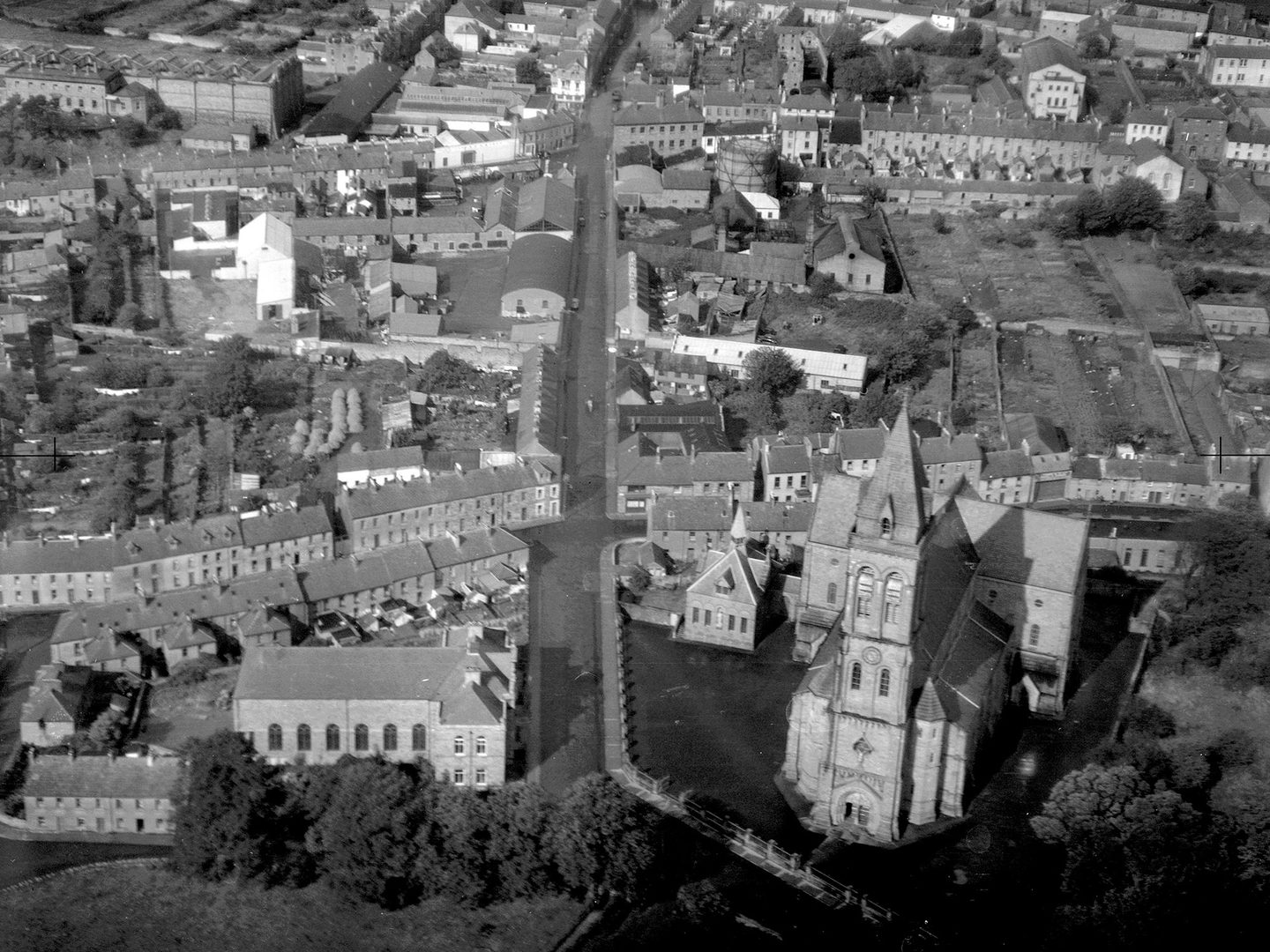 A historic black and white oblique vertical aerial photo of a town. In the foreground is a cathedral with a tower and short transepts.
