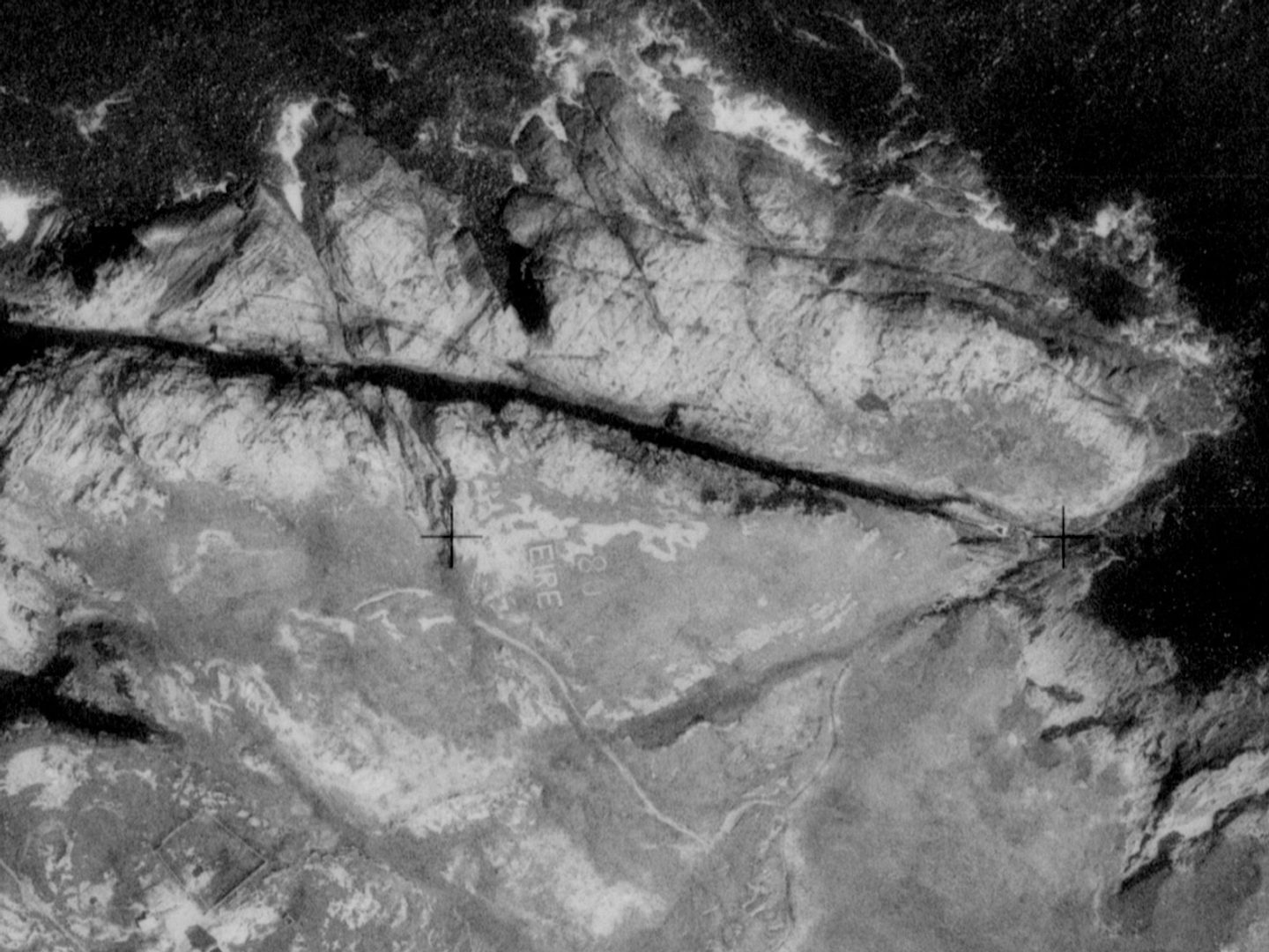 A historic black and white vertical aerial photo of a headland with a ragged, rocky coastline fringed with white waves by a dark sea. The letters '80 EIRE' are faintly picked out in white on the grass of the headland.