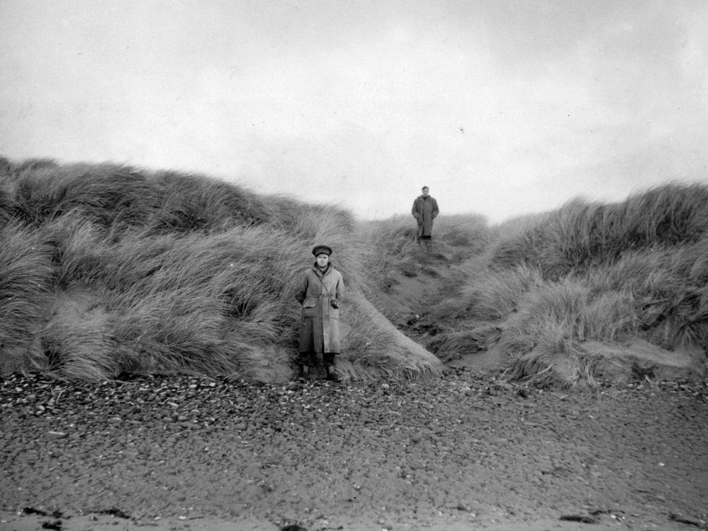 A historic black and white photo of two men in uniform on a beach. The nearer man stands on the sand, with the further away man on the top of a grassy dune.