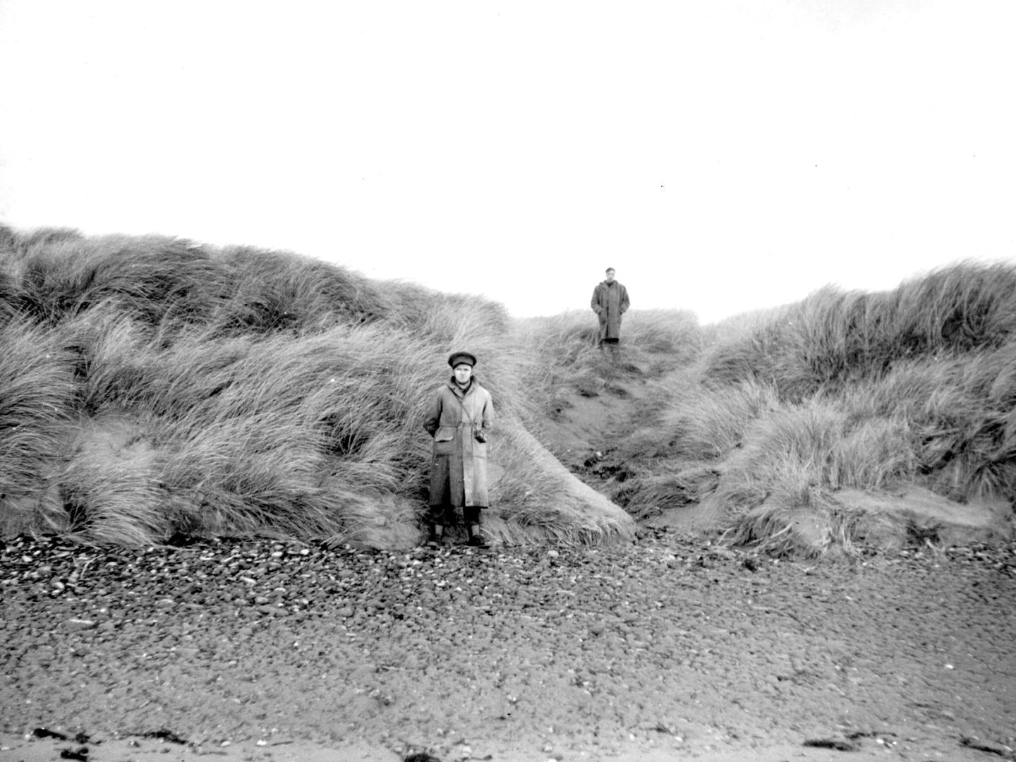 A historic black and white photo of two men in uniform on a beach. The nearer man stands on the sand, with the further away man on the top of a grassy dune.