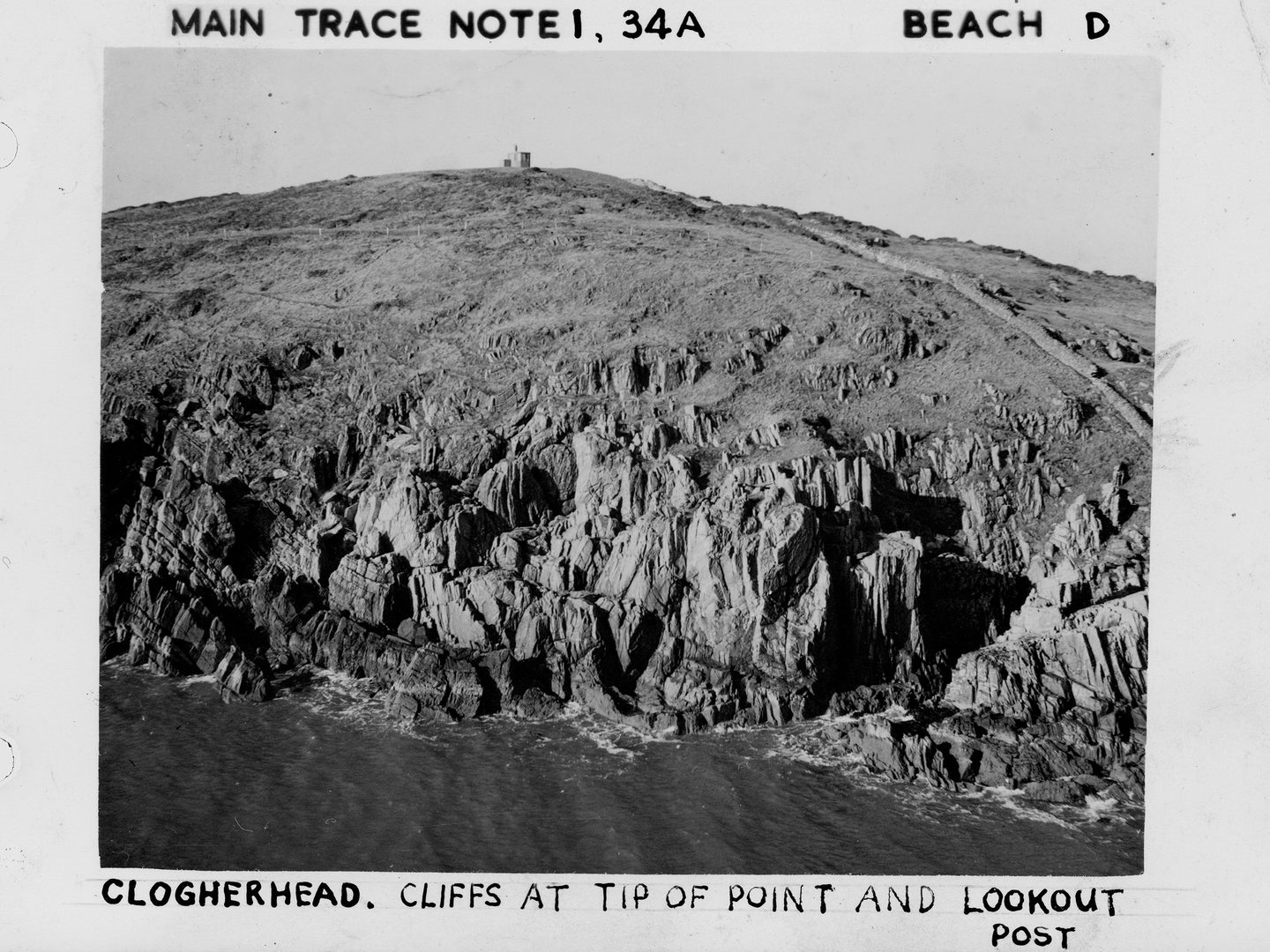 A historic black and white oblique aerial photo of a rocky cliff above a calm sea. Grassy slopes continue to run uphill from top of the cliffs towards a small concrete building.