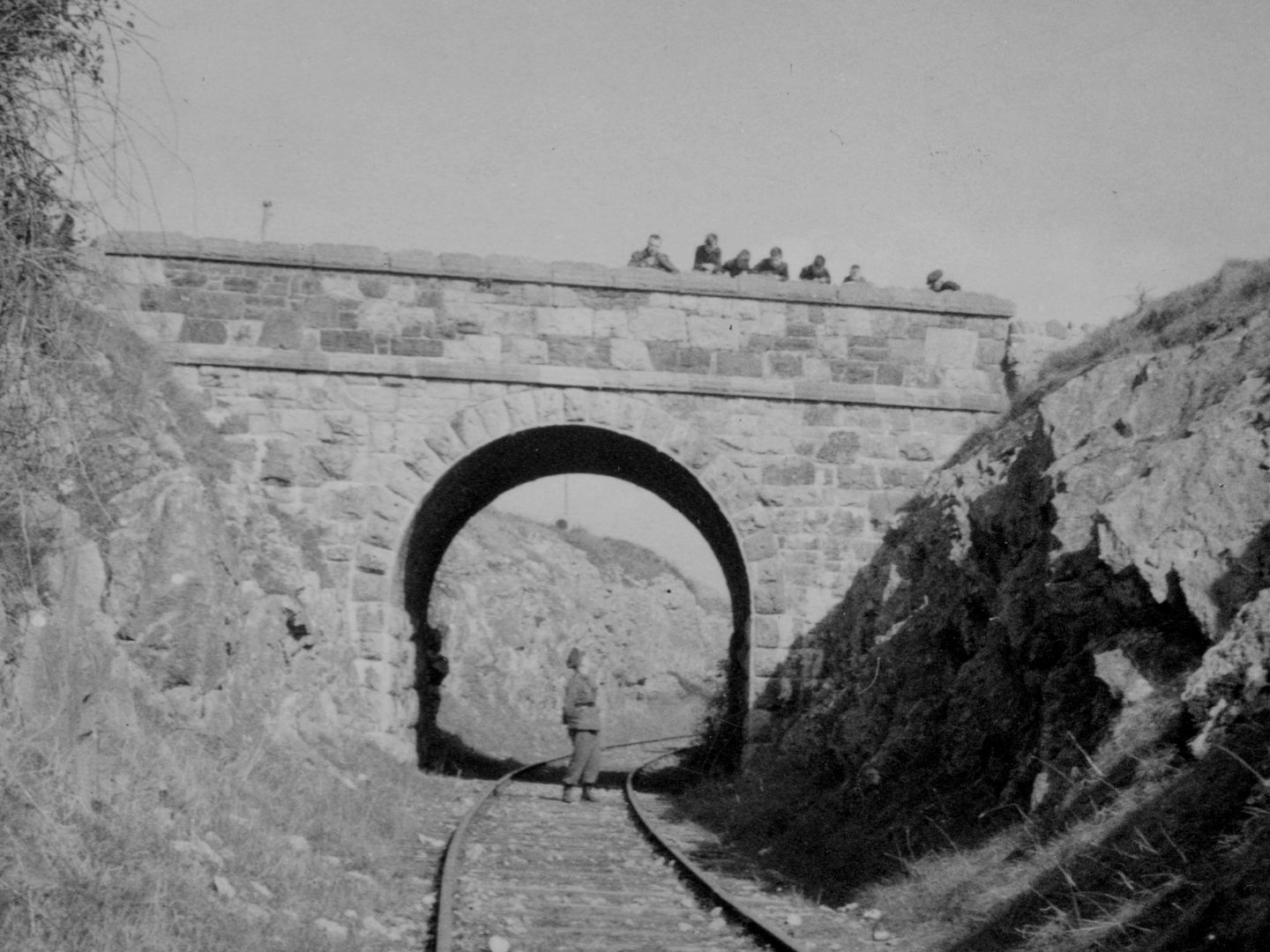 A historic black and white photo of train tracks running under a stone bridge. A man in uniform stands under the bridge, looking up towards a group of people looking down at him over the parapet.