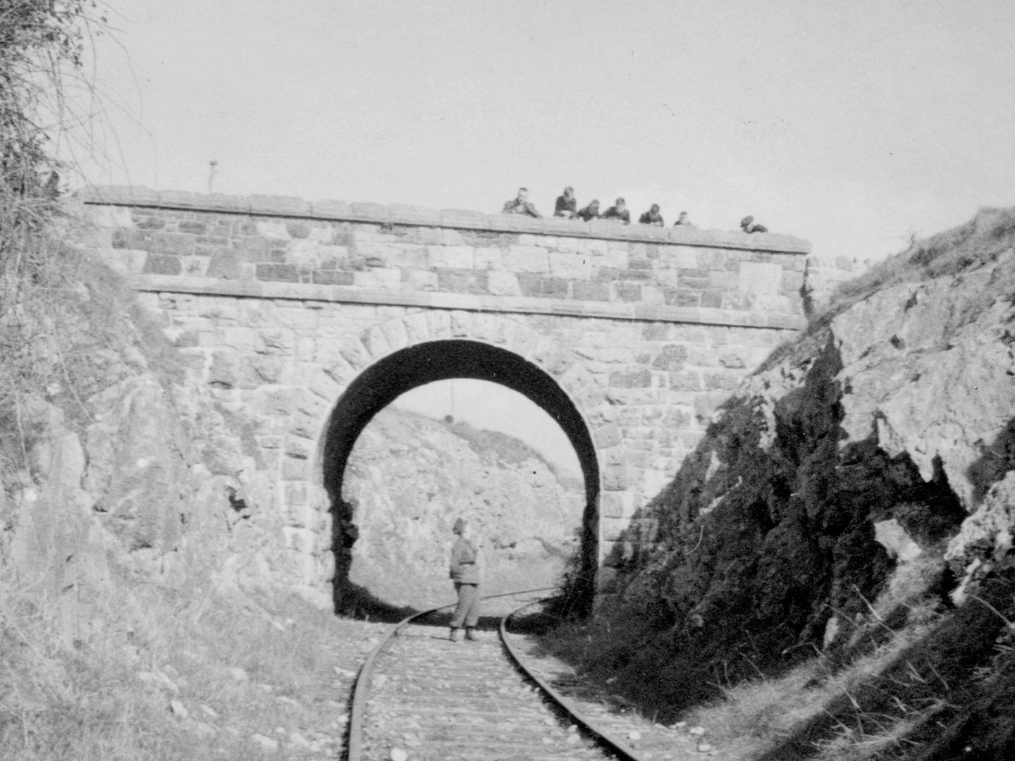 A historic black and white photo of train tracks running under a stone bridge. A man in uniform stands under the bridge, looking up towards a group of people looking down at him over the parapet.
