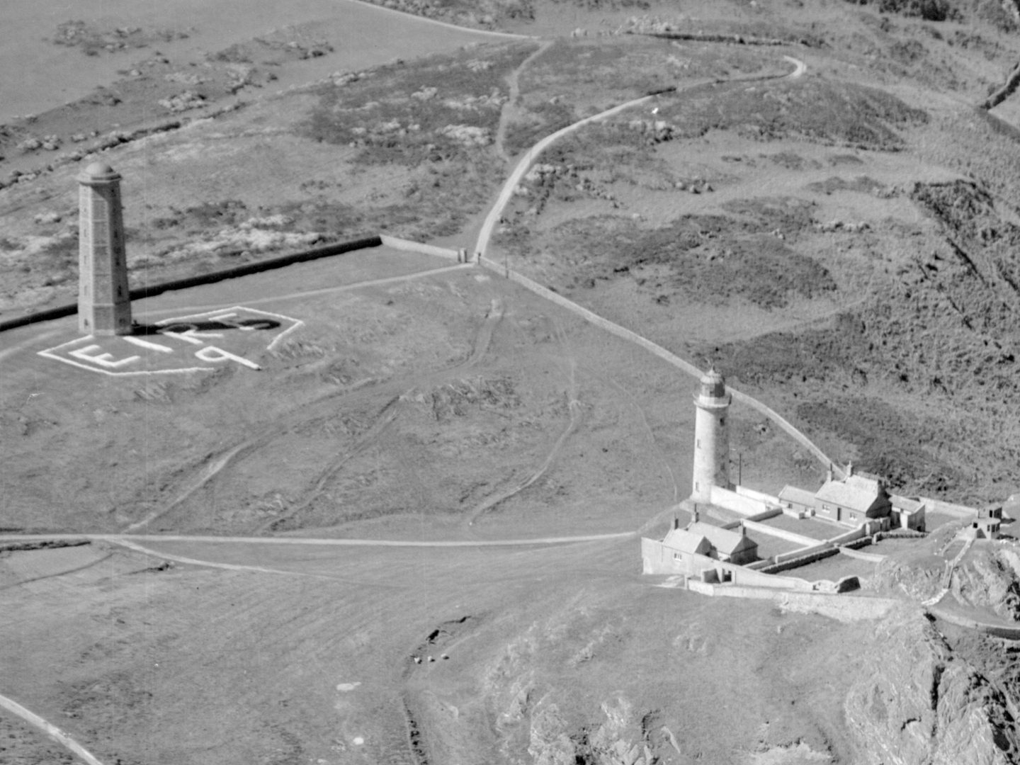 A historic black and white oblique aerial photo of two lighthouses in a grassy landscape. The left-hand lighthouse is grey and multifaceted, while the right-hand lighthouse is shorter, cylindrical and white. The letters 'EIRE 9' have been picked out in white just below the left-hand lighthouse.