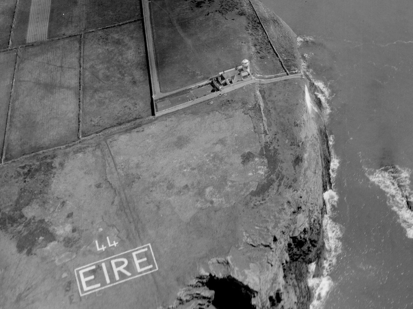 A historic black and white oblique aerial photo of a coastline. In the background are fields divided by walls, with a grassy area in the foreground. To the right cliffs drop into the sea. A lighthouse and associated buildings are just above centre. In the grass in the foreground the letters '44 EIRE' are picked out in white.