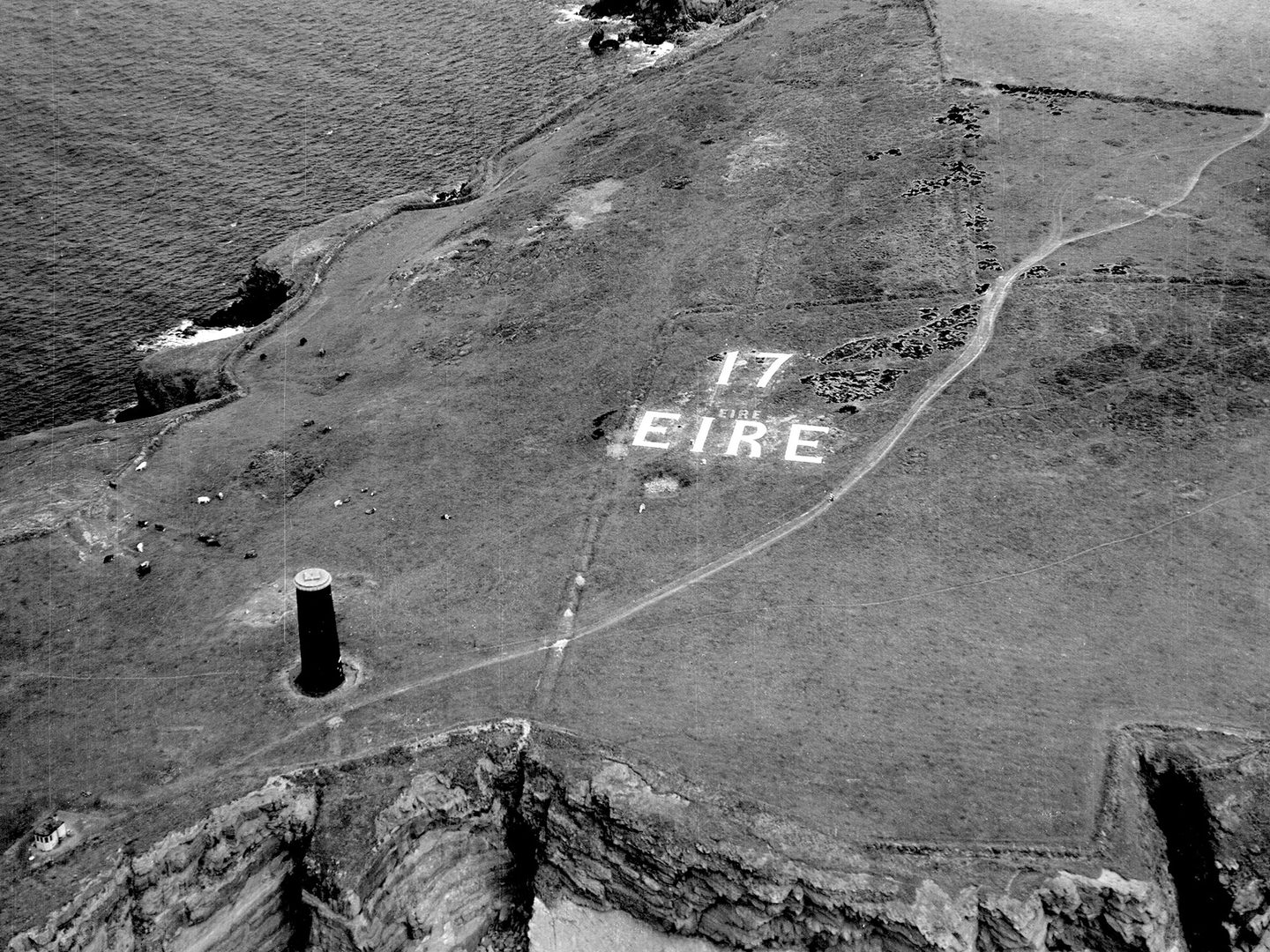 A historic black and white oblique aerial photo of a grassy headland, flanked on both sides by rocky cliffs dropping to the sea. To the left is a cylindrical stone beacon while in the centre of the image the letters '17 EIRE' are picked out in white on the grass.