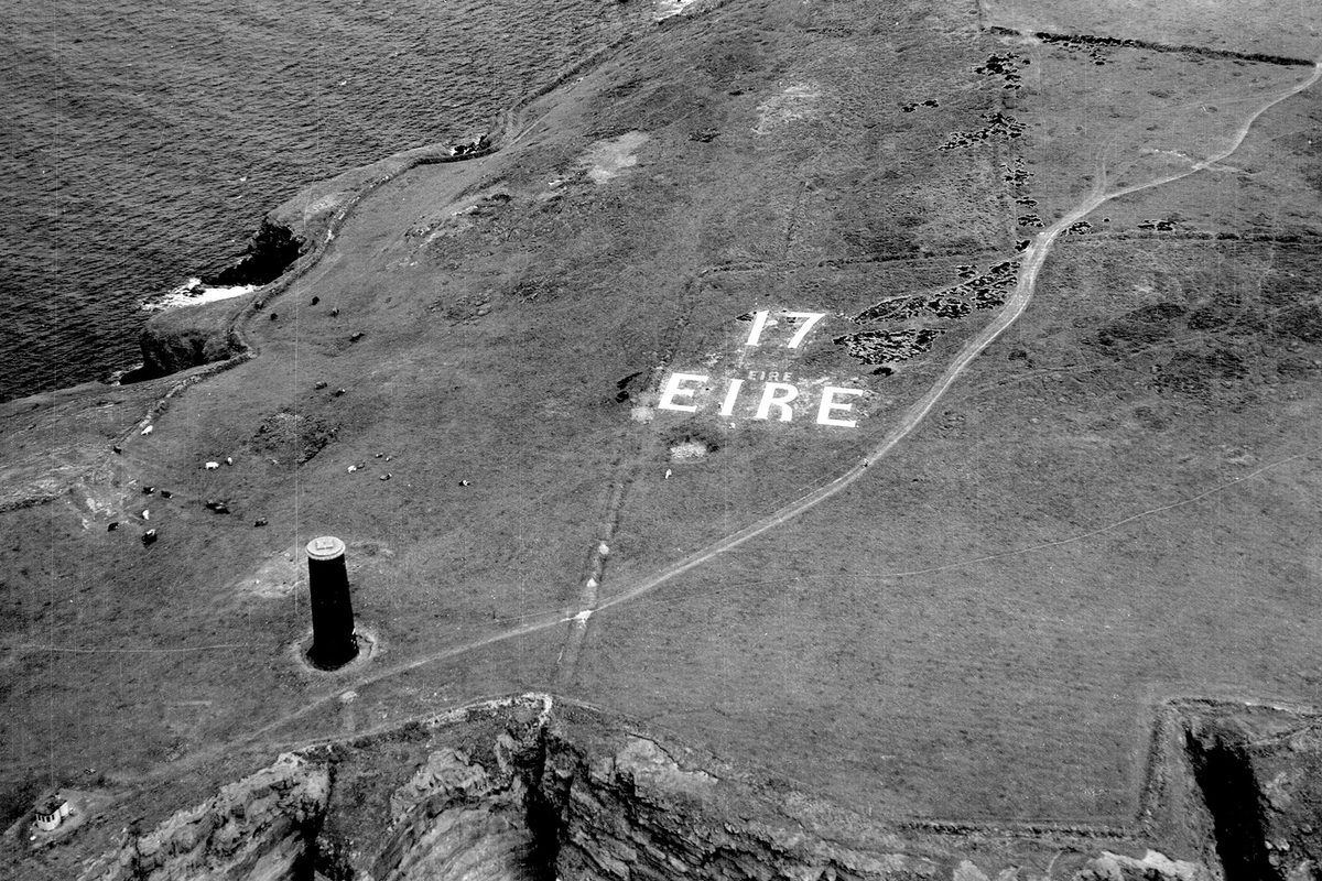 A historic black and white oblique aerial photo of a grassy headland, flanked on both sides by rocky cliffs dropping to the sea. To the left is a cylindrical stone beacon while in the centre of the image the letters '17 EIRE' are picked out in white on the grass.