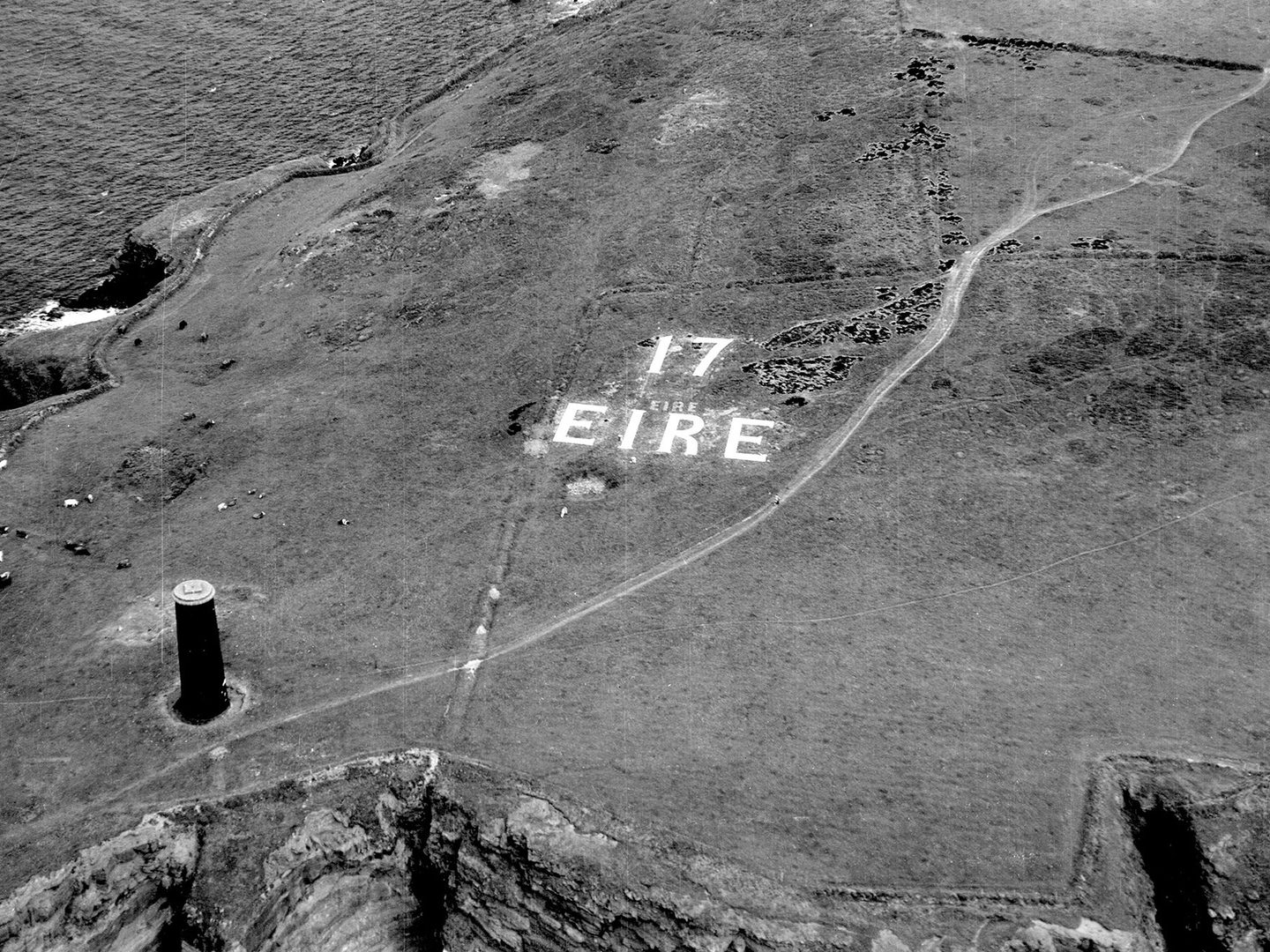 A historic black and white oblique aerial photo of a grassy headland, flanked on both sides by rocky cliffs dropping to the sea. To the left is a cylindrical stone beacon while in the centre of the image the letters '17 EIRE' are picked out in white on the grass.