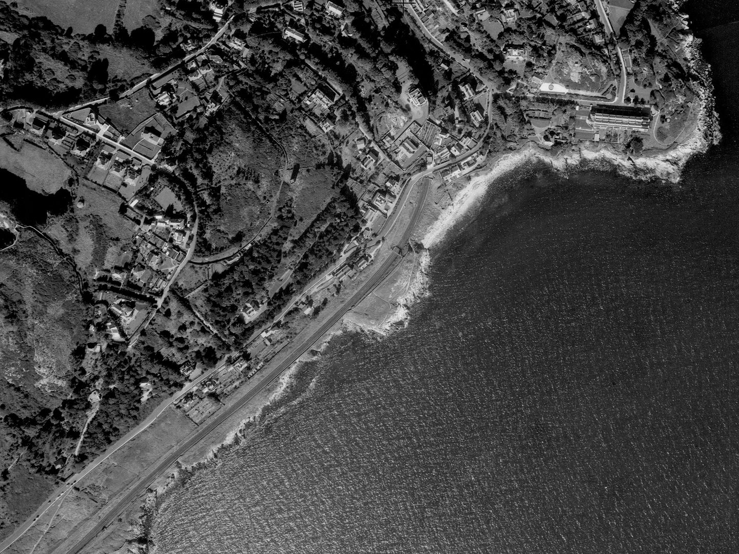 A historic black and white vertical aerial photo of a town on a shoreline, with the sea to the bottom left. A prominent field right on the coast shows no indication of an EIRE Sign.