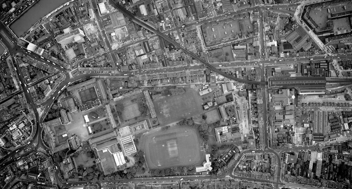 A historic black and white vertical aerial photo of the city of Dublin. To the left are several groups of buildings arranged around courtyards. They form the shape of a Greek cross. To their left are several playing fields.