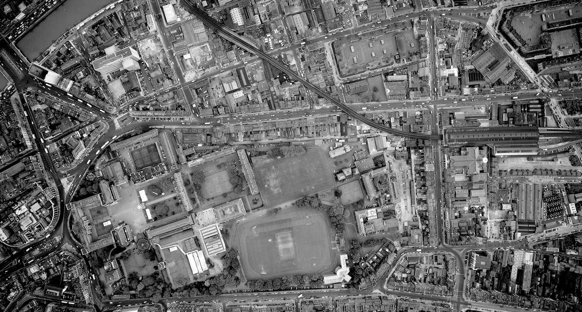 A historic black and white vertical aerial photo of the city of Dublin. To the left are several groups of buildings arranged around courtyards. They form the shape of a Greek cross. To their left are several playing fields.
