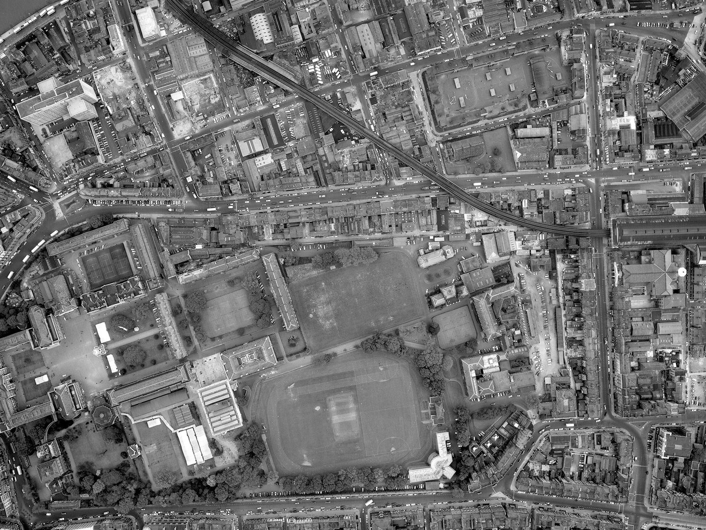 A historic black and white vertical aerial photo of the city of Dublin. To the left are several groups of buildings arranged around courtyards. They form the shape of a Greek cross. To their left are several playing fields.
