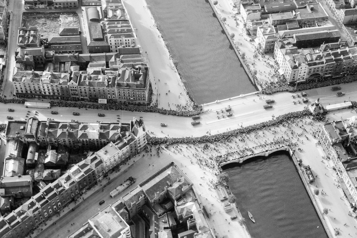 A historic black and white oblique aerial photo of a parade of military vehicles along a building-lined, broad street. The street is lined with large crowds and passes over a river running from the top of the image to the bottom towards the right-hand side.