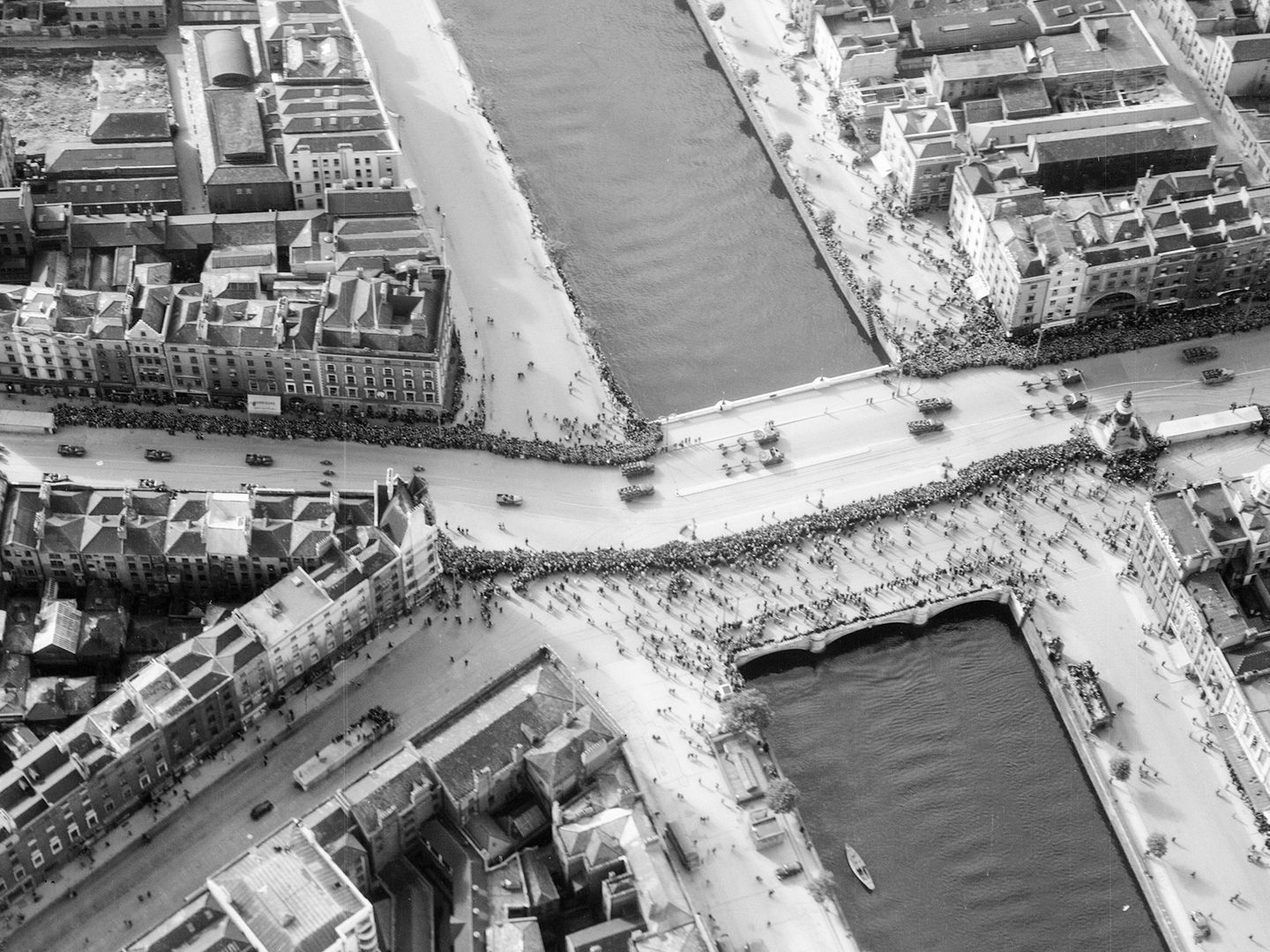 A historic black and white oblique aerial photo of a parade of military vehicles along a building-lined, broad street. The street is lined with large crowds and passes over a river running from the top of the image to the bottom towards the right-hand side.