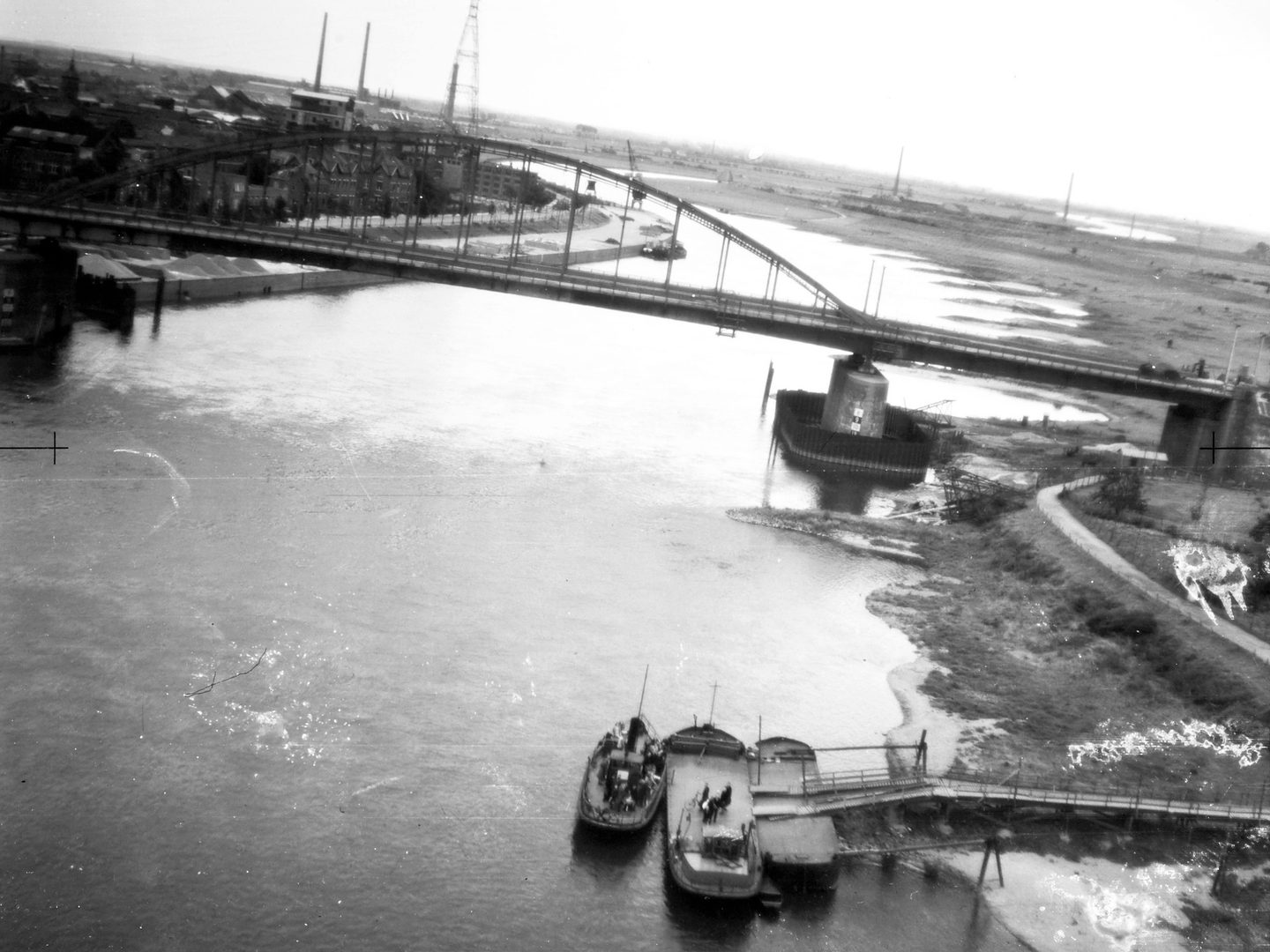 A black and white oblique aerial photo of a river. In the foreground pedestrians about to board a boat look up at the camera. Beyond them is a bridge suspended from curved iron trusses. Industrial buildings and pylons are visible in the background.