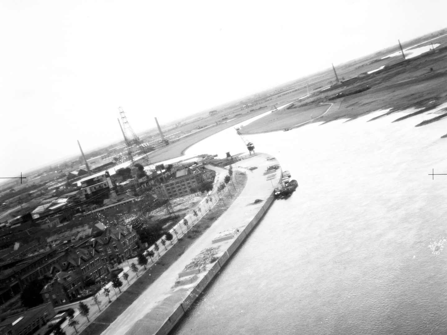A black and white oblique aerial photo of a river, taken with the plane at an angle so the horizon is tilted from lower left to upper right. Industrial buildings and pylons are visible on the left bank. The right bank is flat and dark coloured.