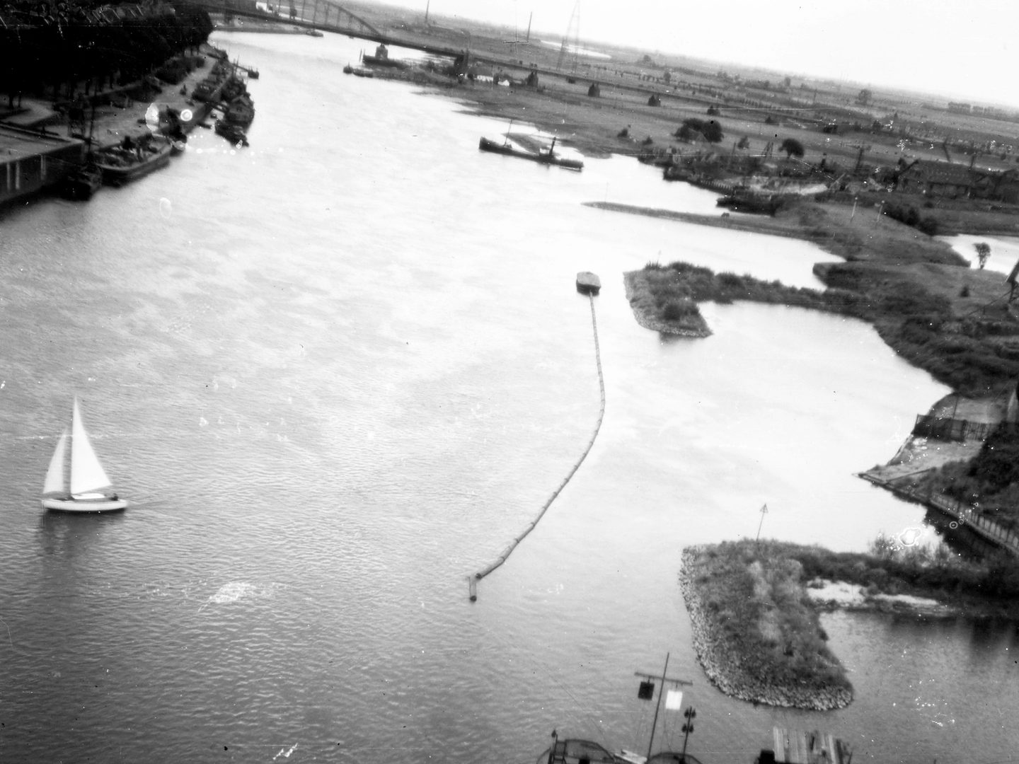 A black and white oblique aerial photo of a river, taken with the plane at an angle so the horizon tilts from upper left to lower right. A boat with white triangular sails is bottom left. A bridge with curved iron trusses is in the distance.