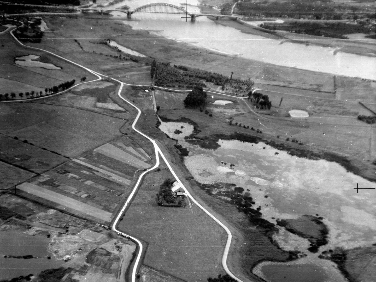 A black and white oblique aerial photo of a landscape, with a network of white roads crossing marshy ground. A few trees are visible, with a house just below centre. There is a curving river top right crossed by a bridge with 5 spans.