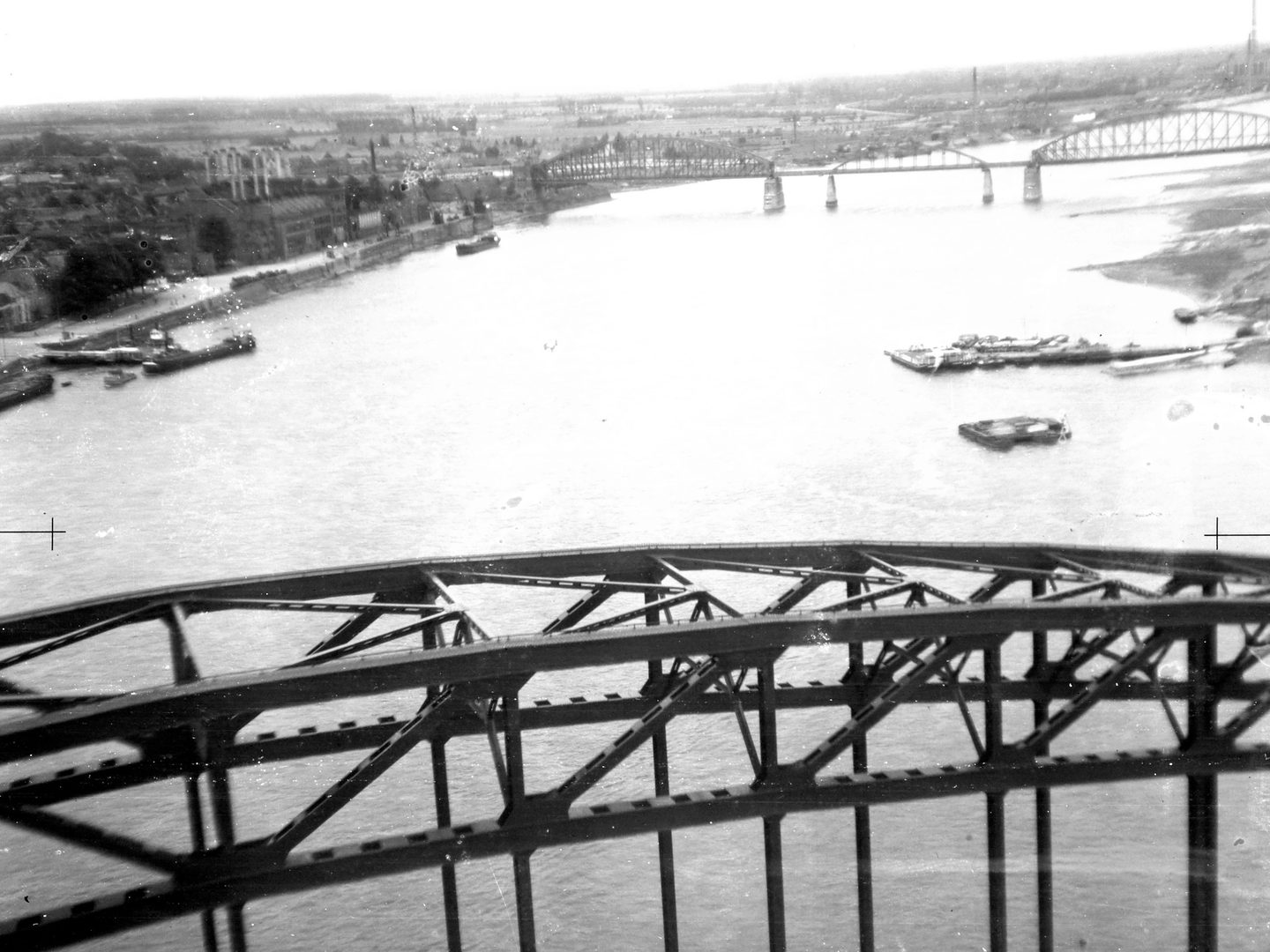 A black and white oblique aerial photo of a river. A network of steel girders fills the bottom third of the image. Boats are visible beyond the bridge. In the distance are industrial buildings on the left riverbank and a second bridge with 3 spans.