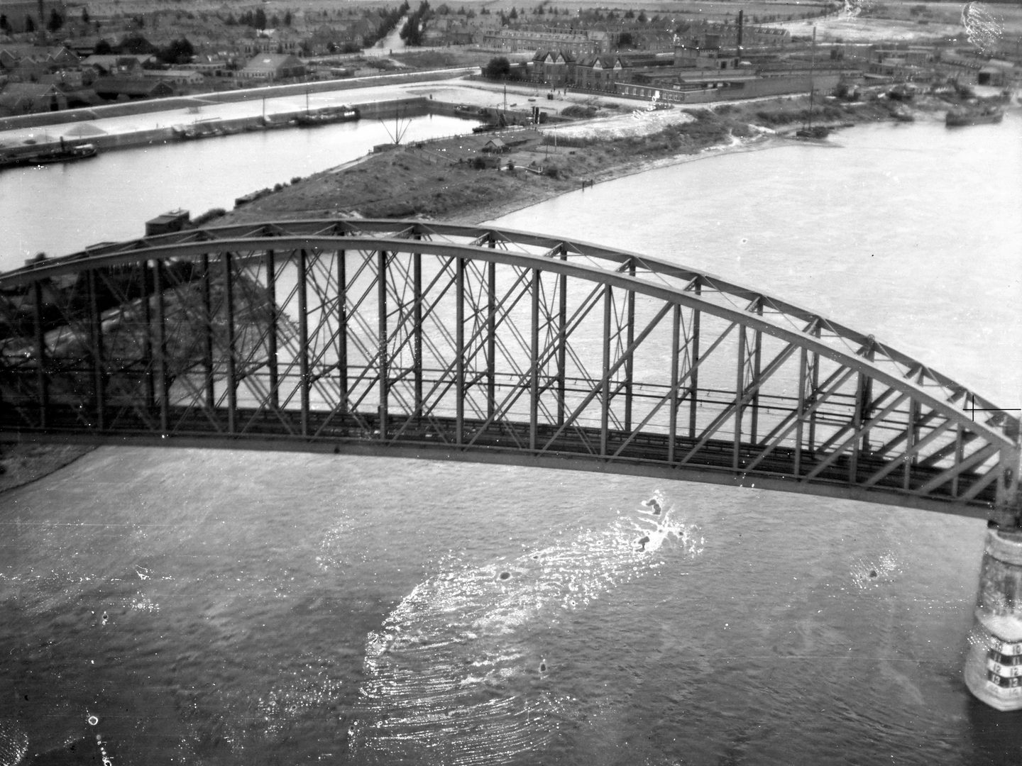 A black and white oblique aerial photo of a bridge across a river. The bridge is suspended from a curved iron truss and a network of girders. Only the left bank of the river is visible. On it are houses and industrial buildings with chimneys.
