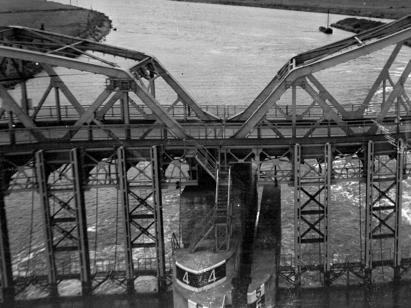A black and white oblique aerial photo of a bridge across a river. The bridge rests on a large concrete pier. Iron structures either side of the pier further support the bridge, which is itself largely made of iron. A single boat is visible beyond.