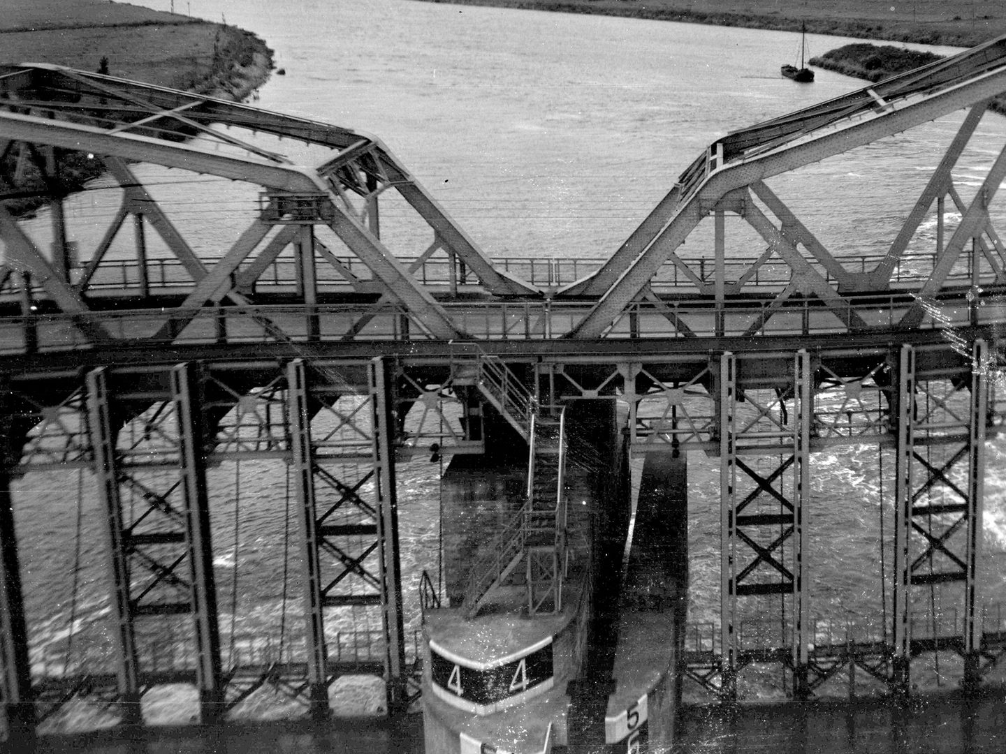 A black and white oblique aerial photo of a bridge across a river. The bridge rests on a large concrete pier. Iron structures either side of the pier further support the bridge, which is itself largely made of iron. A single boat is visible beyond.