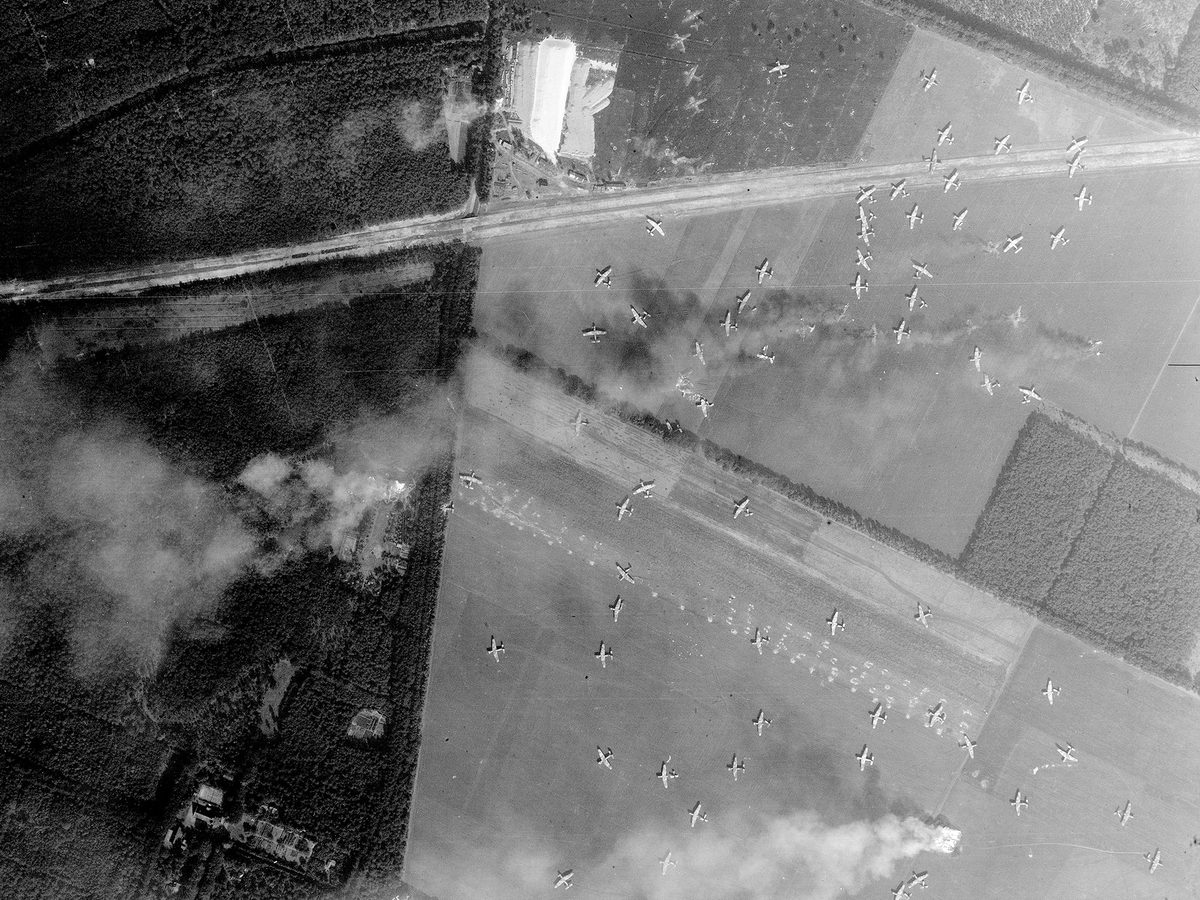 A black and white aerial photo of a landscape. Woods fill the left-hand third of the image, there are mostly fields to the right and a road cuts across the top half. Scattered across the fields are over 60 gliders. Smoke blows from right to left.