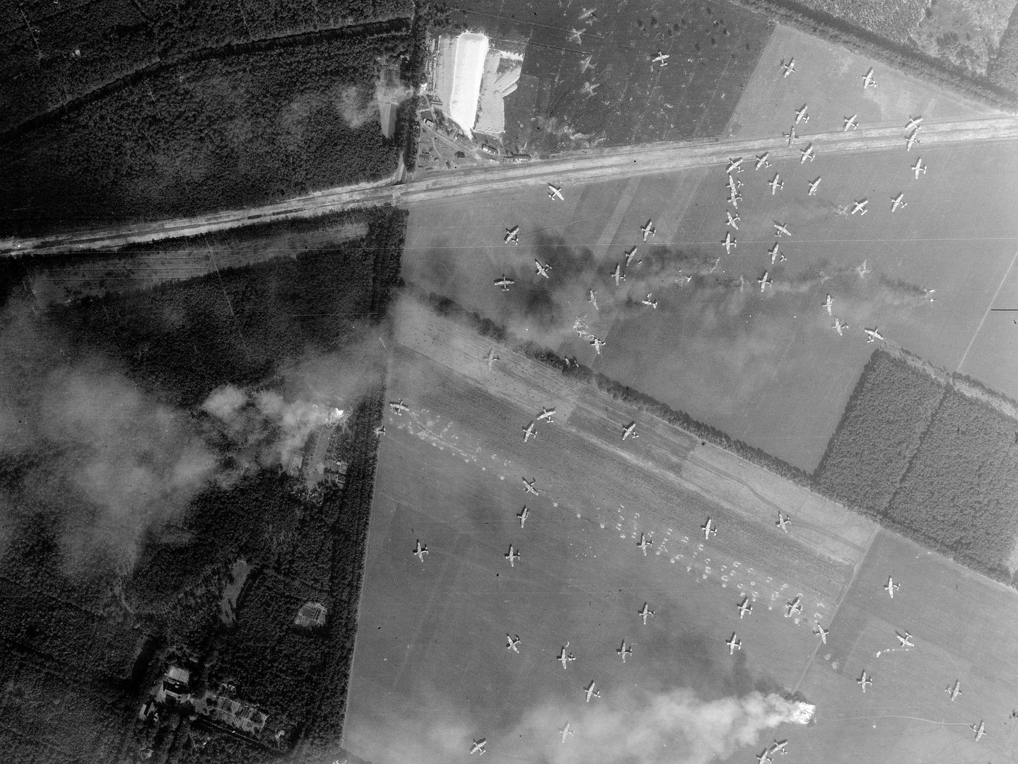 A black and white aerial photo of a landscape. Woods fill the left-hand third of the image, there are mostly fields to the right and a road cuts across the top half. Scattered across the fields are over 60 gliders. Smoke blows from right to left.