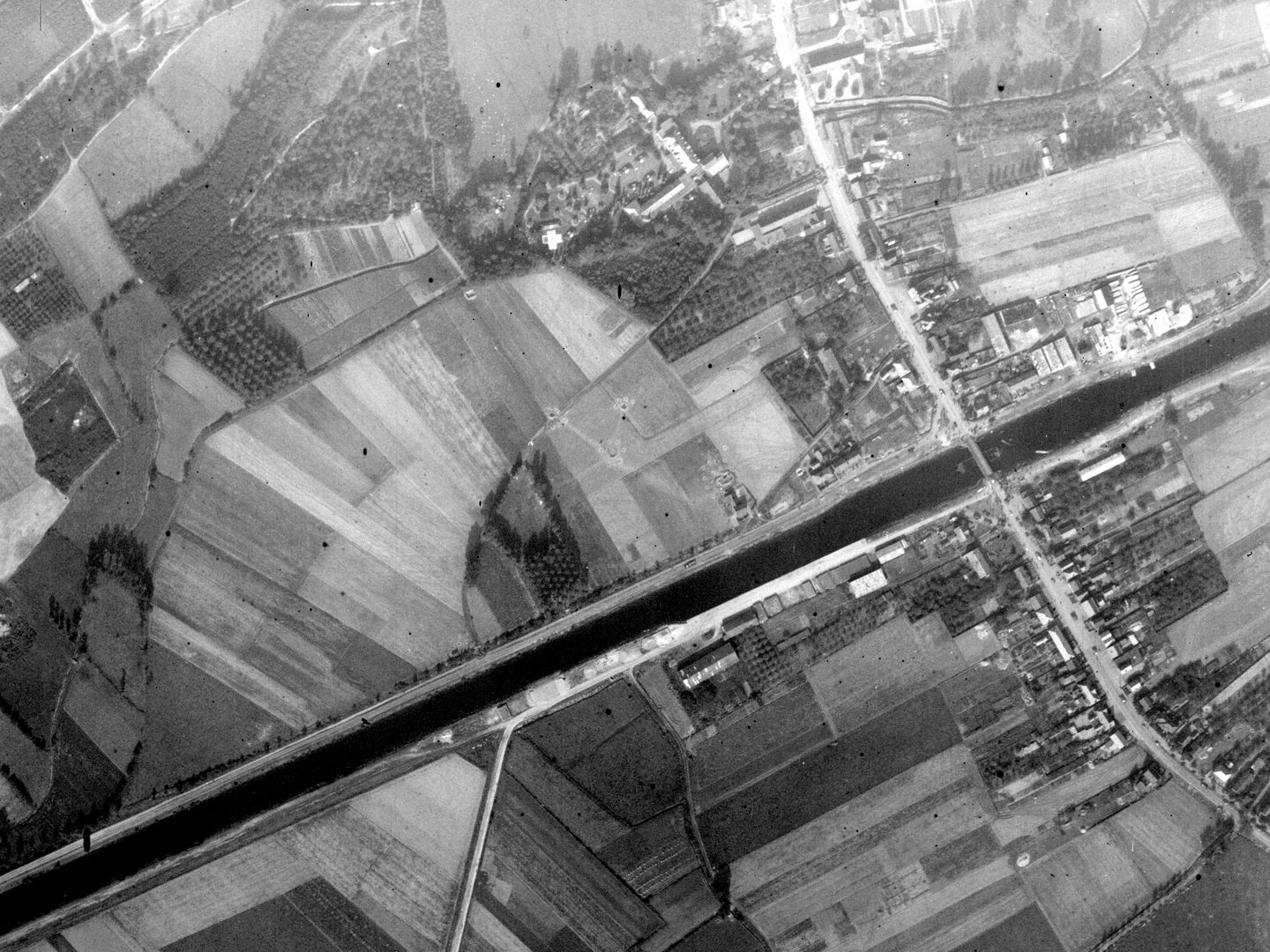 A black and white aerial photo of a canal stretching from the bottom left corner to near the top right. The canal is surrounded by woods and fields. There is a town at the right side of the image, straddling the canal with a road running through it.