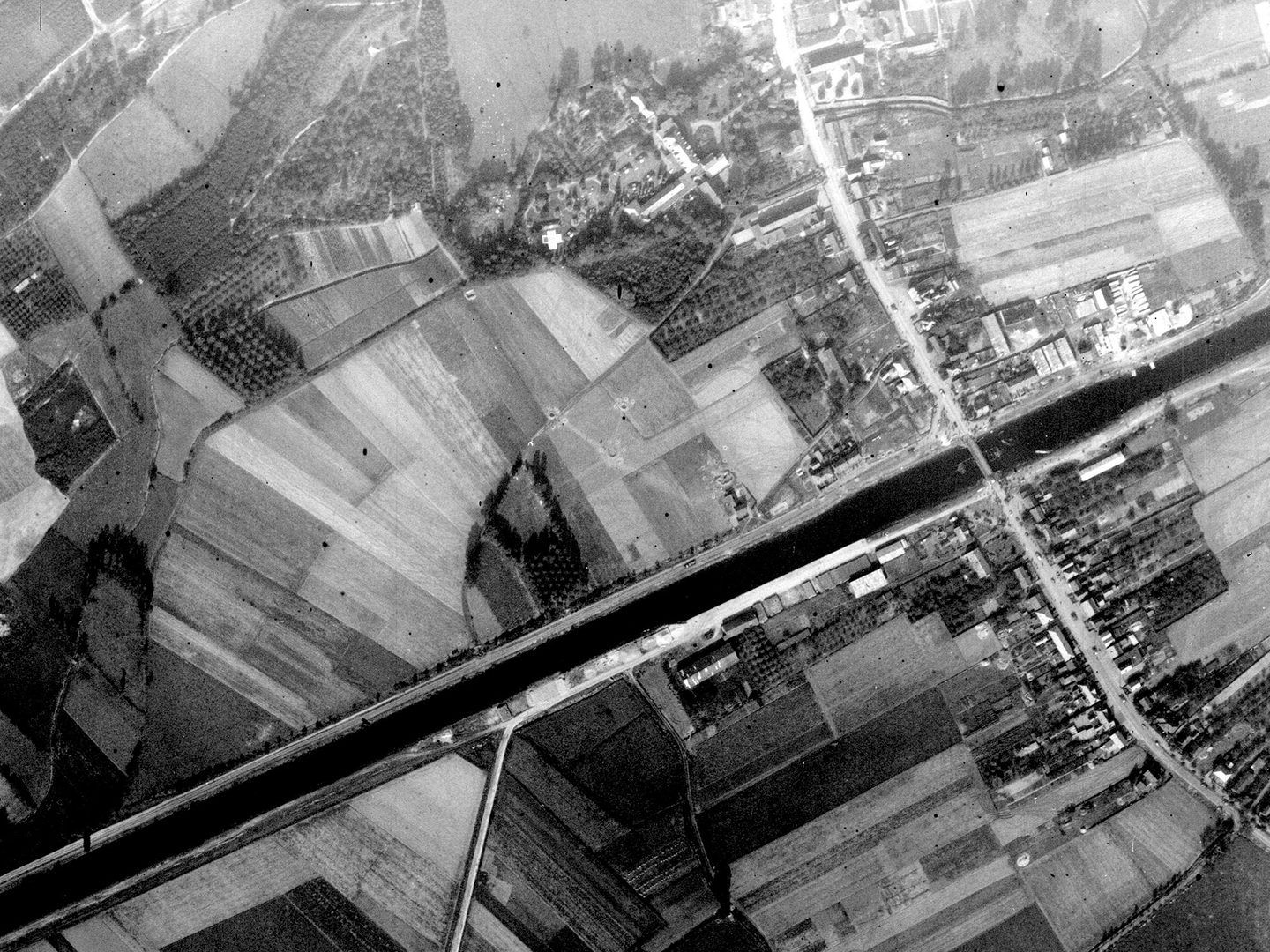A black and white aerial photo of a canal stretching from the bottom left corner to near the top right. The canal is surrounded by woods and fields. There is a town at the right side of the image, straddling the canal with a road running through it.