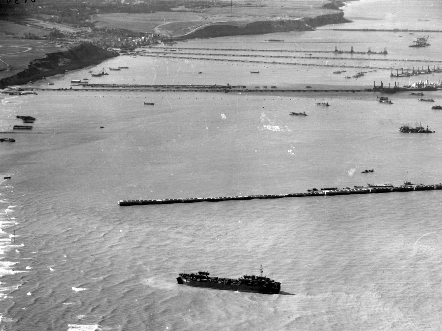 A black and white oblique aerial photo of a coastline. Several temporary piers have been constructed to protrude from the coastline, and are interspersed with numerous naval vessels.
