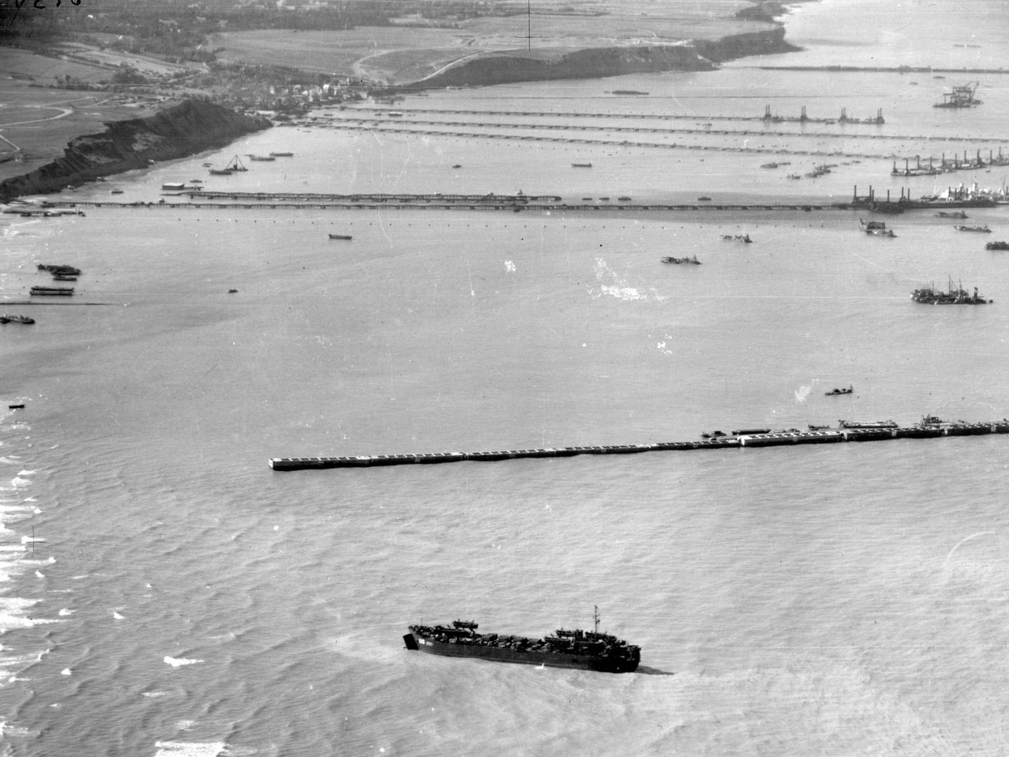A black and white oblique aerial photo of a coastline. Several temporary piers have been constructed to protrude from the coastline, and are interspersed with numerous naval vessels.