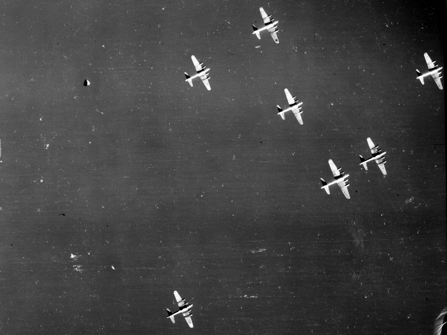 A black and white aerial photo of 7 B-17 bombers flying in formation. The planes are silver in colour and stand out against the dark background of the image. 6 of the planes are close together in the top right, 1 is by itself bottom left.