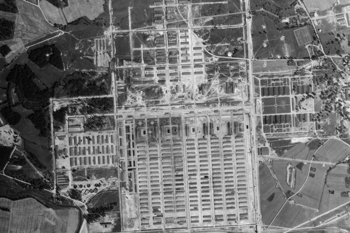 A black and white aerial photo of the Auschwitz-Birkenau extermination camp. Huts are arranged in grids in different sections around the camp, which is surrounded by woods and fields.