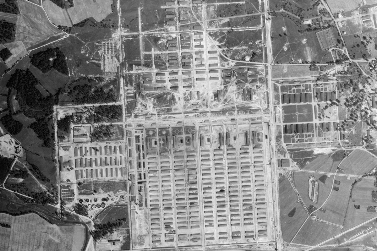 A black and white aerial photo of the Auschwitz-Birkenau extermination camp. Huts are arranged in grids in different sections around the camp, which is surrounded by woods and fields.
