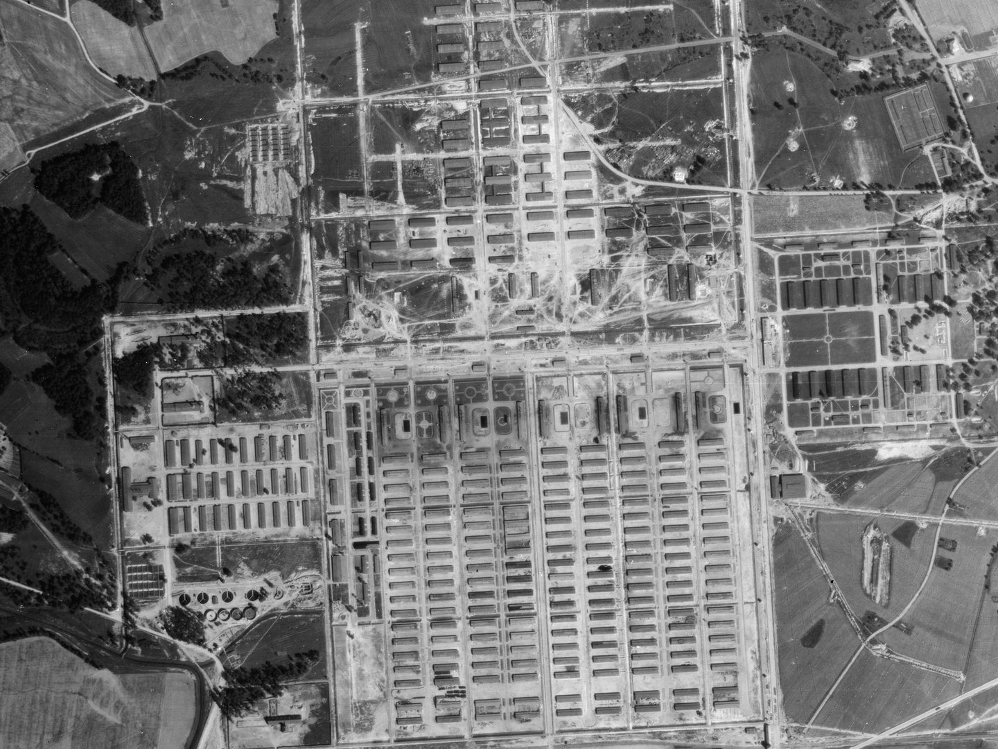 A black and white aerial photo of the Auschwitz-Birkenau extermination camp. Huts are arranged in grids in different sections around the camp, which is surrounded by woods and fields.