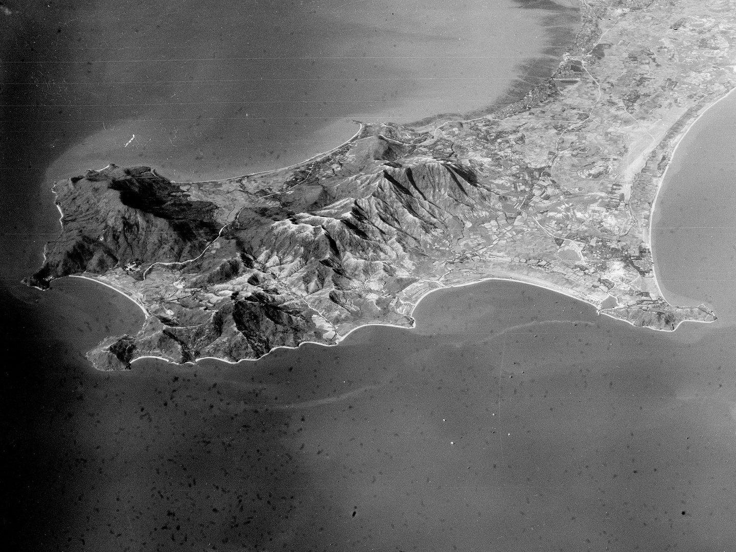A historic black and white oblique aerial photo of a mountainous peninsula stretching from the top right corner towards the bottom left, surrounded by sea on 3 sides.