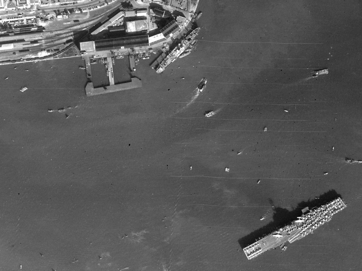 A black and white aerial photo of the sea, with numerous ships on it. Most prominent is a large aircraft carrier carrying many planes at the bottom right corner. A small area of land with industrial buildings is visible at the top left corner.