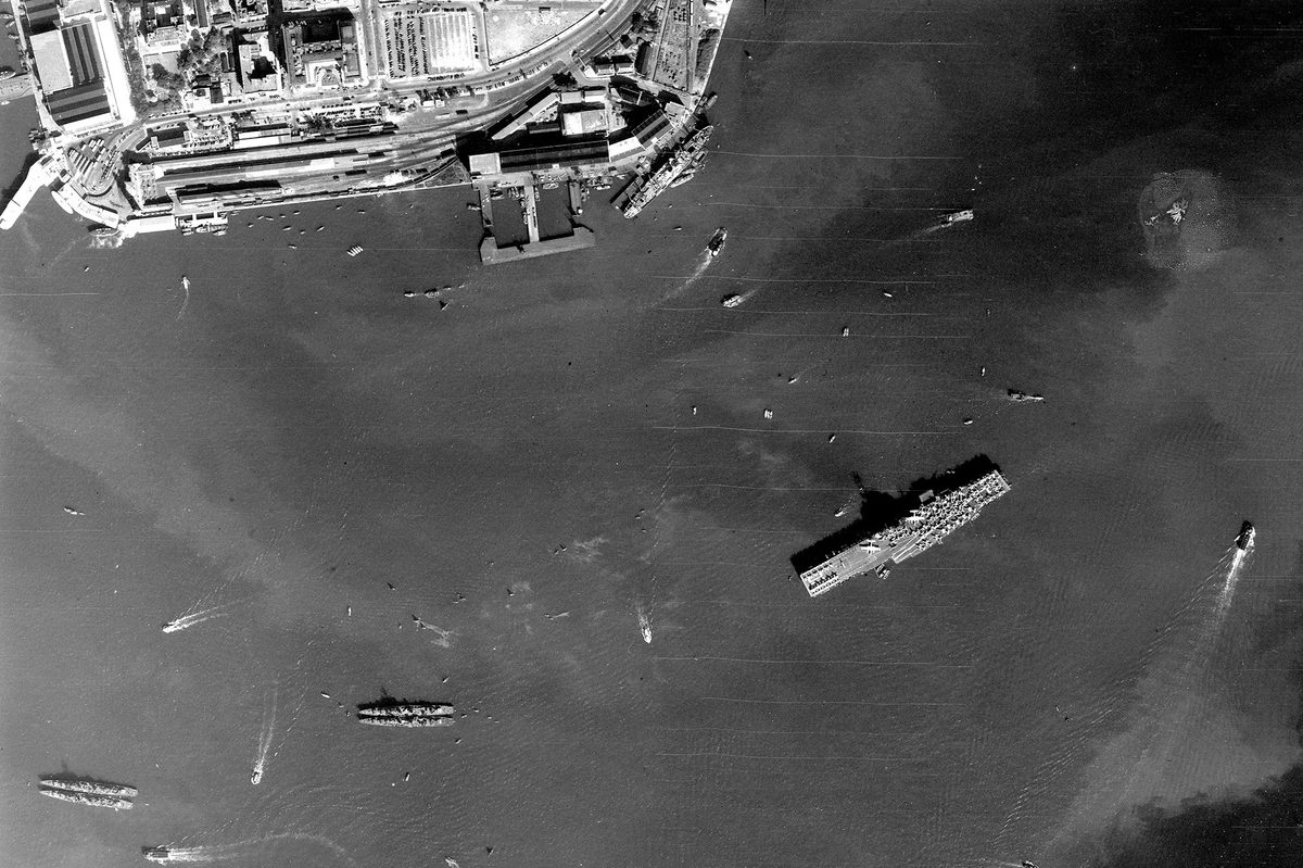 A historic black and white vertical aerial image of a large aircraft carrier, its deck covered with aircraft. Several other vessels are visible on the sea around the aircraft carrier. Docks and a port are visible on the edge of a small portion of land at the top edge of the image.