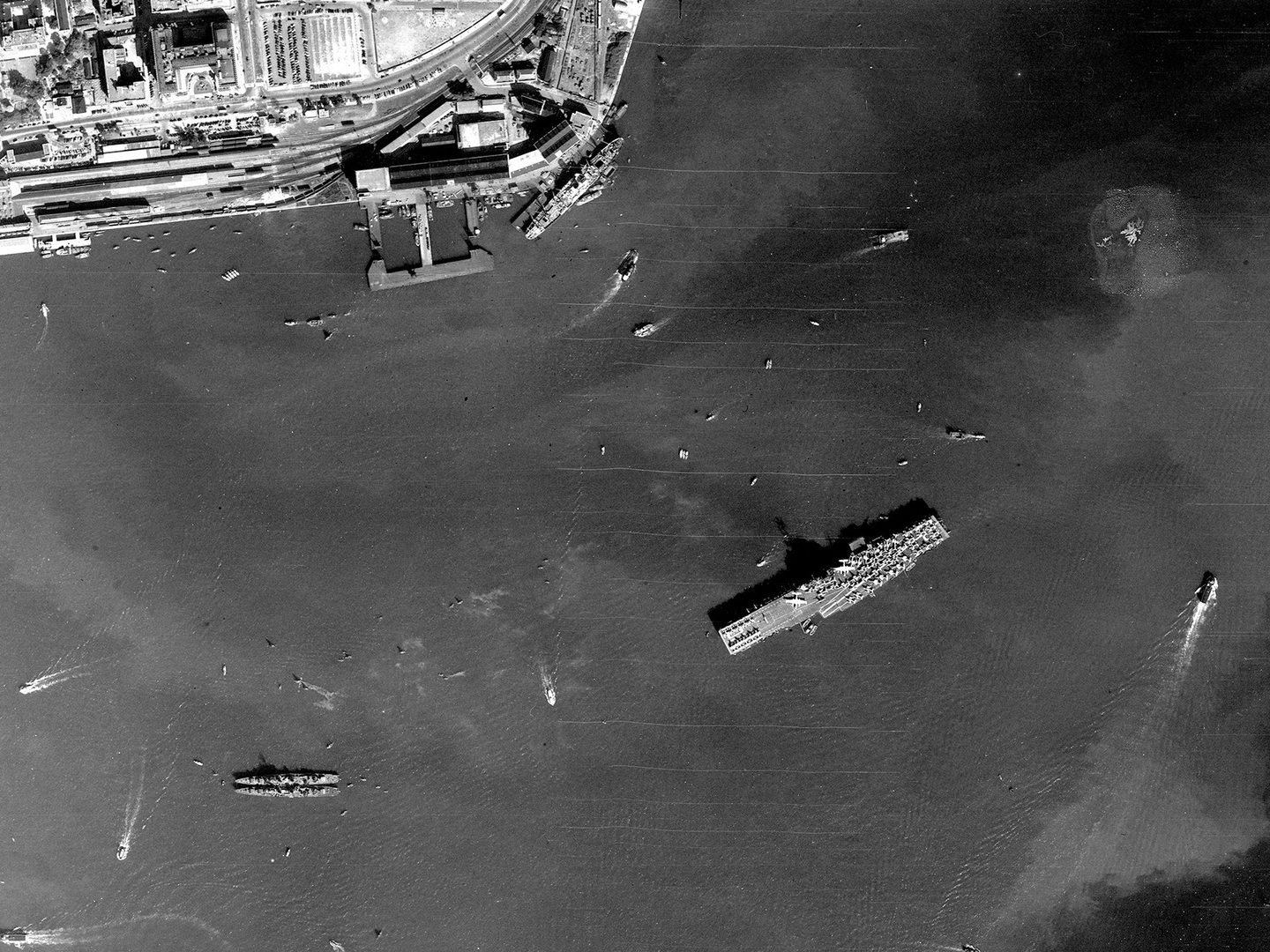 A historic black and white vertical aerial image of a large aircraft carrier, its deck covered with aircraft. Several other vessels are visible on the sea around the aircraft carrier. Docks and a port are visible on the edge of a small portion of land at the top edge of the image.