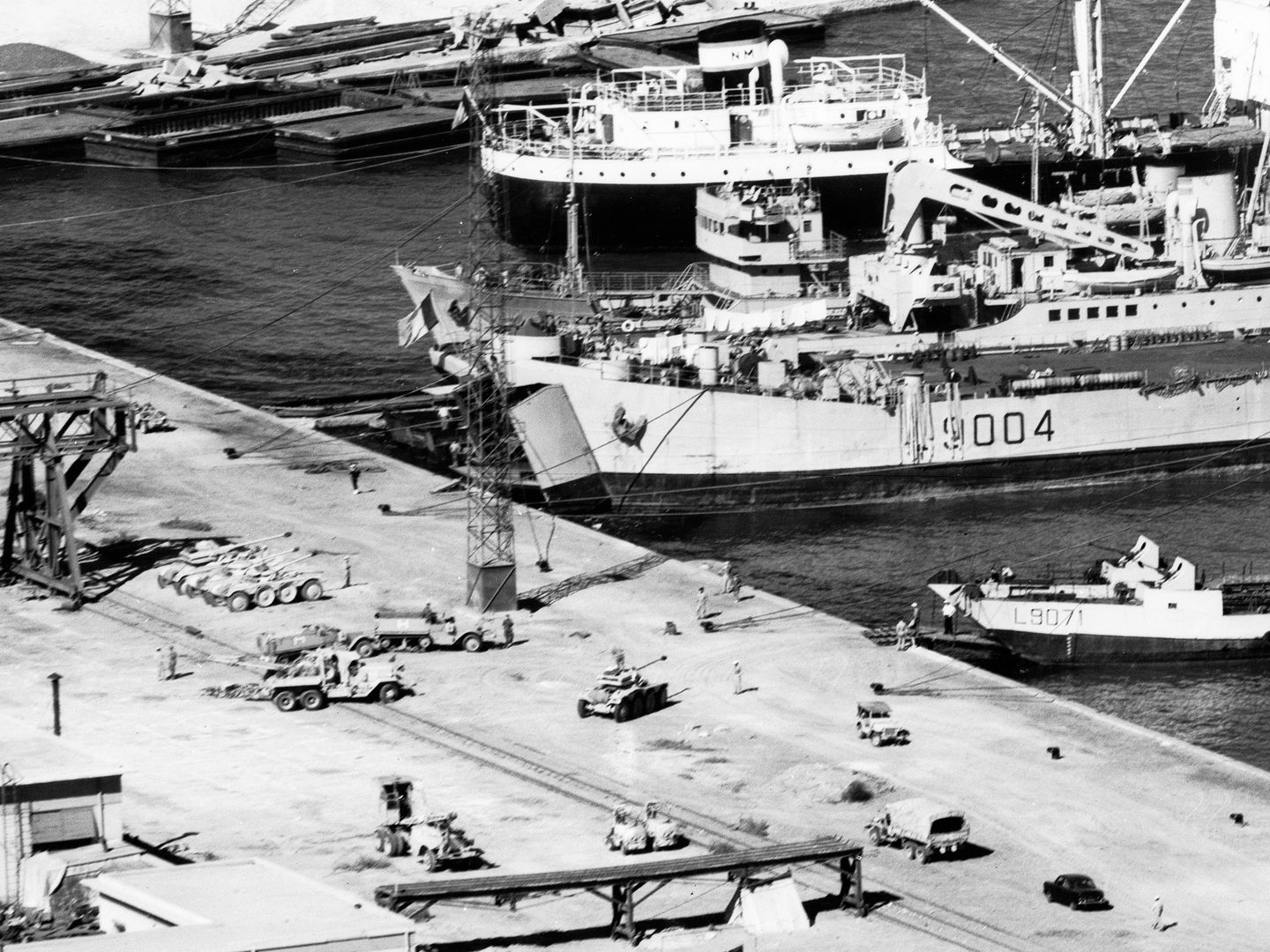 A historic black and white vertical aerial photo of a dockside with vessels drawn up at it. The largest ship has its bow doors open. Various military vehicles are drawn up on the dock ready to be loaded into the ship.