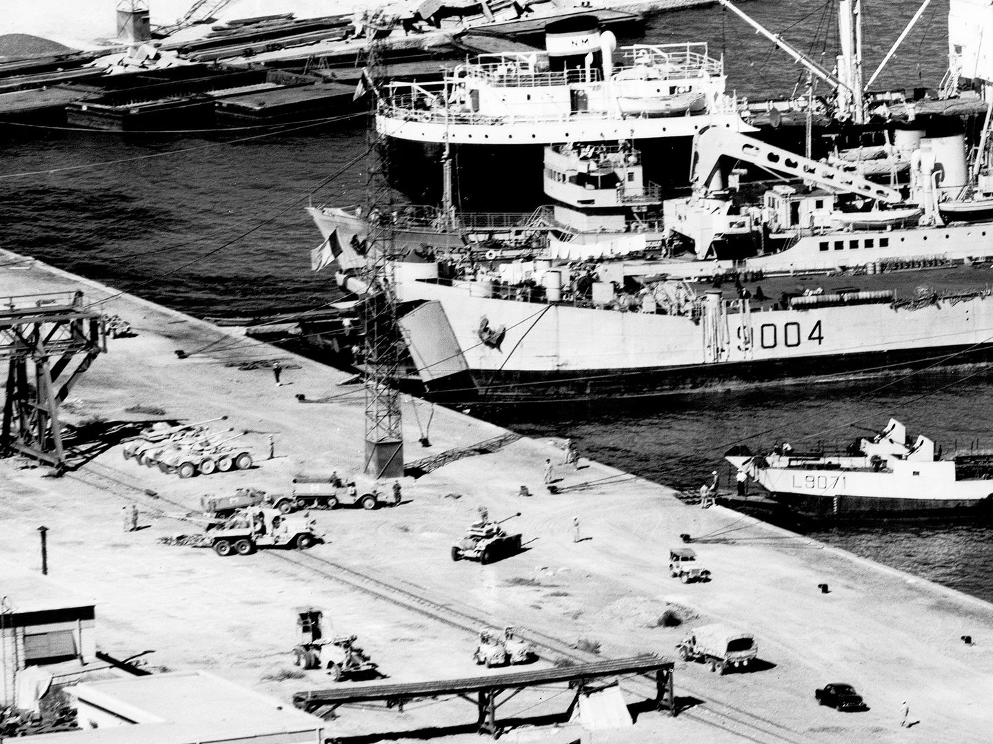 A historic black and white vertical aerial photo of a dockside with vessels drawn up at it. The largest ship has its bow doors open. Various military vehicles are drawn up on the dock ready to be loaded into the ship.