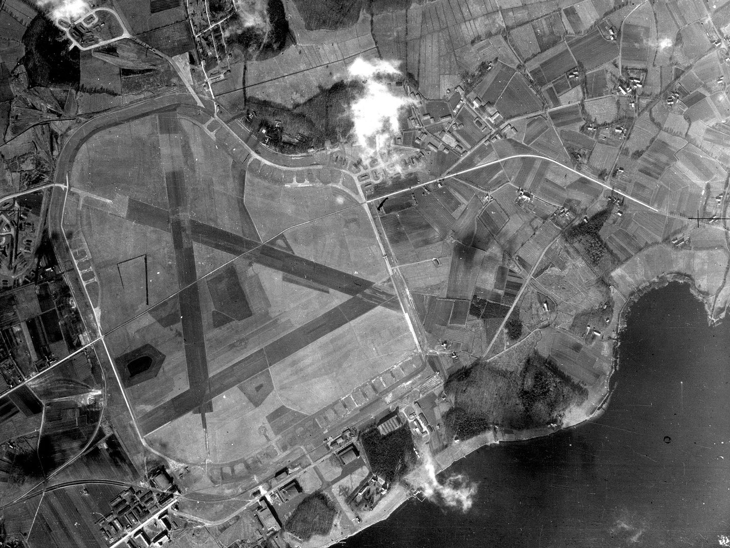 A historic black and white vertical aerial photo of an airfield. 3 runways cross it in an A shape. Fields and buildings are visible around its perimeter. An area of lake occupies the bottom right corner.