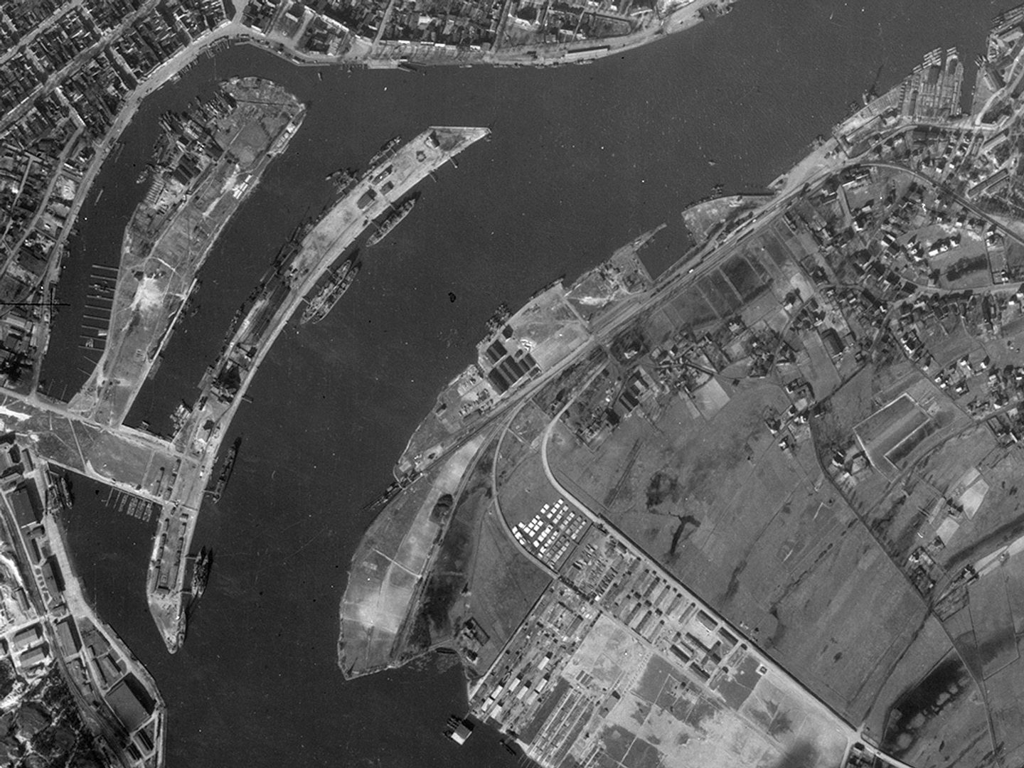 A historic black and white vertical aerial photo of a port on a river. The main course of the river runs from top to bottom of the image, with two smaller branches to the left. Numerous vessels are on the river and pulled up at the docks.