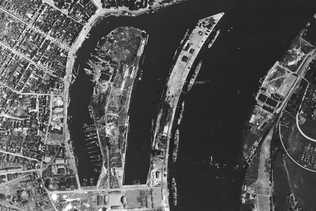 A historic black and white vertical aerial photo of a port on a river. The main course of the river runs from top to bottom of the image, with two smaller branches to the left. Numerous vessels are on the river and pulled up at the docks.