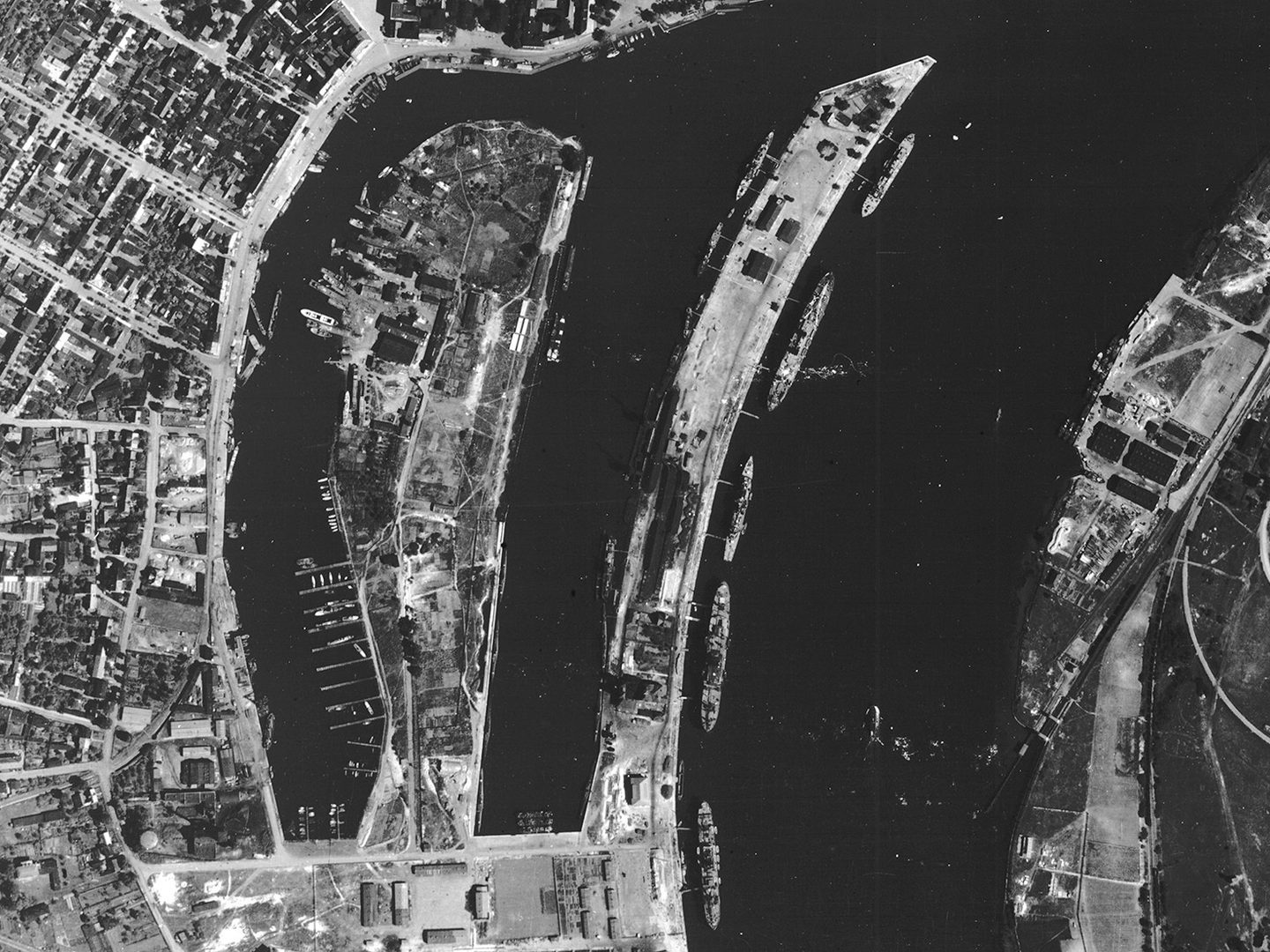 A historic black and white vertical aerial photo of a port on a river. The main course of the river runs from top to bottom of the image, with two smaller branches to the left. Numerous vessels are on the river and pulled up at the docks.