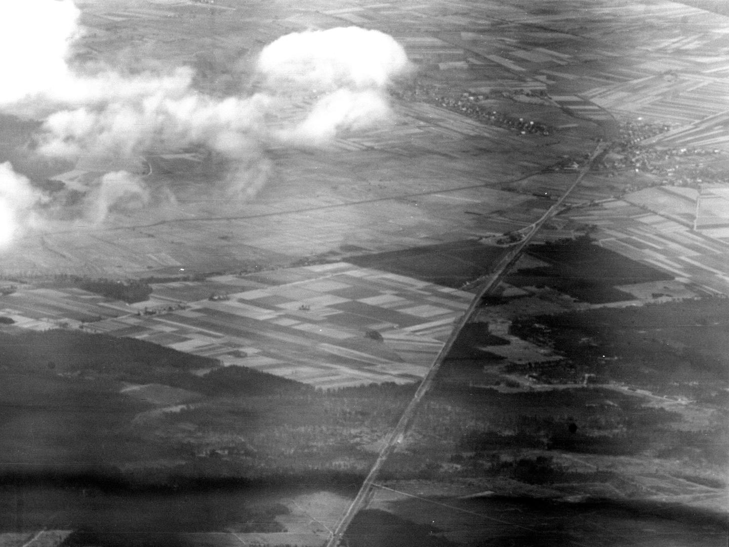 A black and white oblique aerial photo of a landscape with a forest in the foreground and grids of fields stretching into the distance. A road runs through the landscape to a small town in the top right corner. Clouds obscure the top left corner.