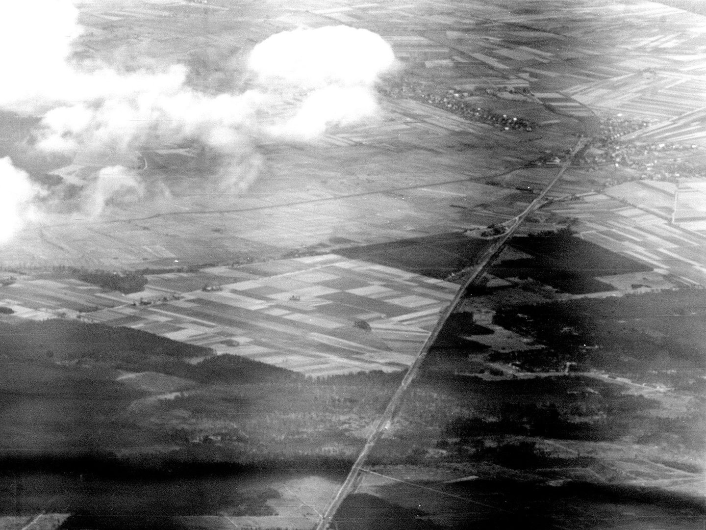 A black and white oblique aerial photo of a landscape with a forest in the foreground and grids of fields stretching into the distance. A road runs through the landscape to a small town in the top right corner. Clouds obscure the top left corner.