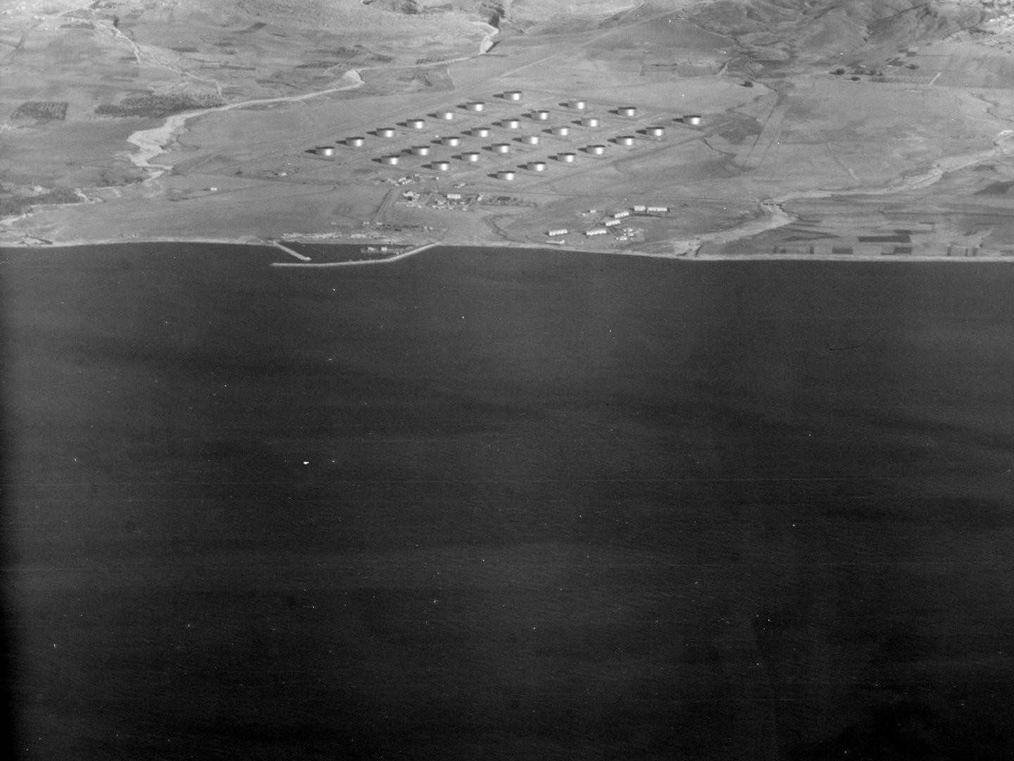 A black and white oblique aerial photo, with a large area of dark sea across the bottom two thirds. A pale-coloured shore is visible in the top third, with 28 shiny circular tanks arranged in a grid.